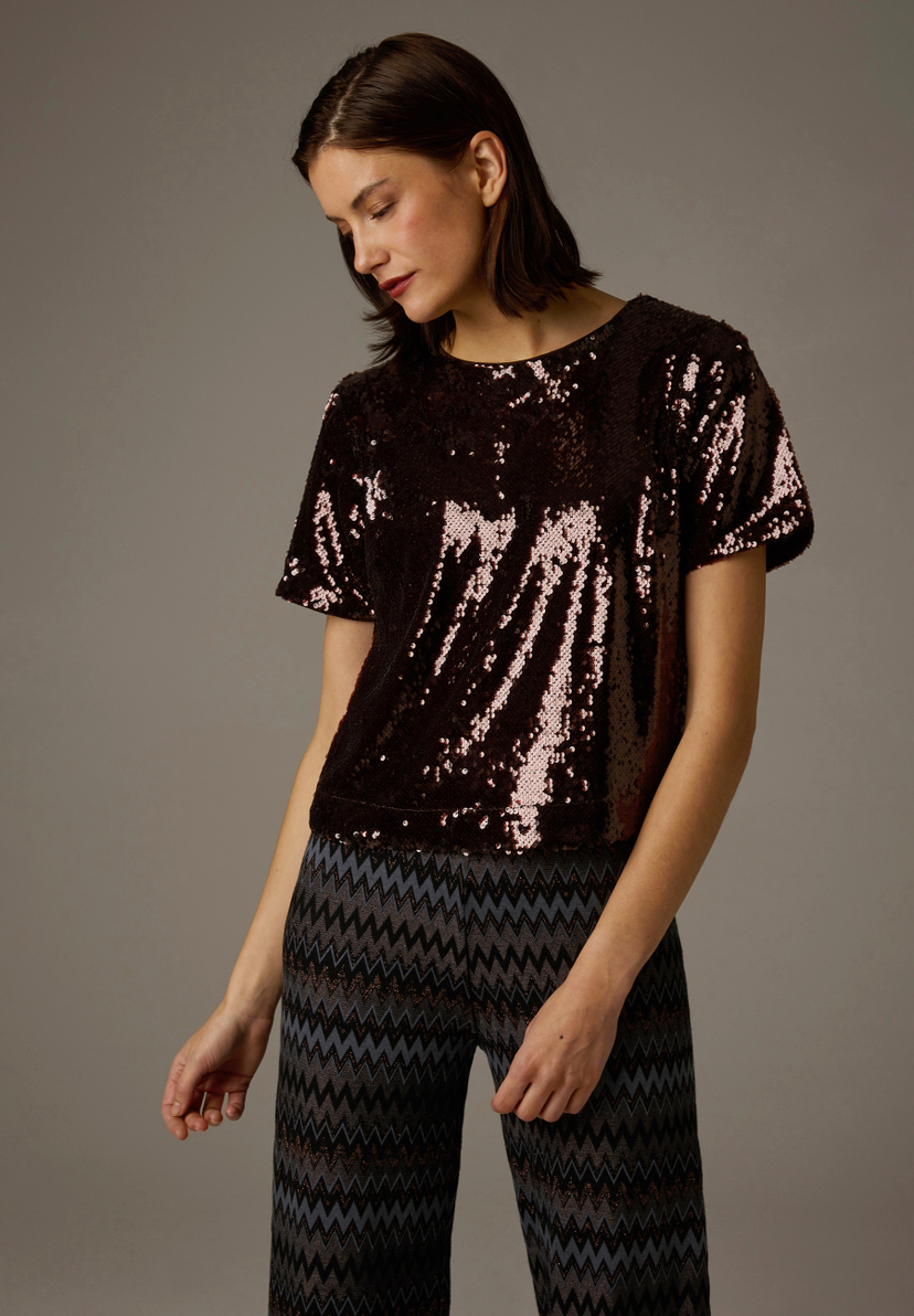 Eine Frau mit verschränkten Armen trägt ein glänzendes MORE & MORE Oberteil aus Polyester mit Pailletten. Eine Frau mit verschränkten Armen trägt ein glänzendes MORE & MORE Oberteil aus Polyester mit Pailletten.