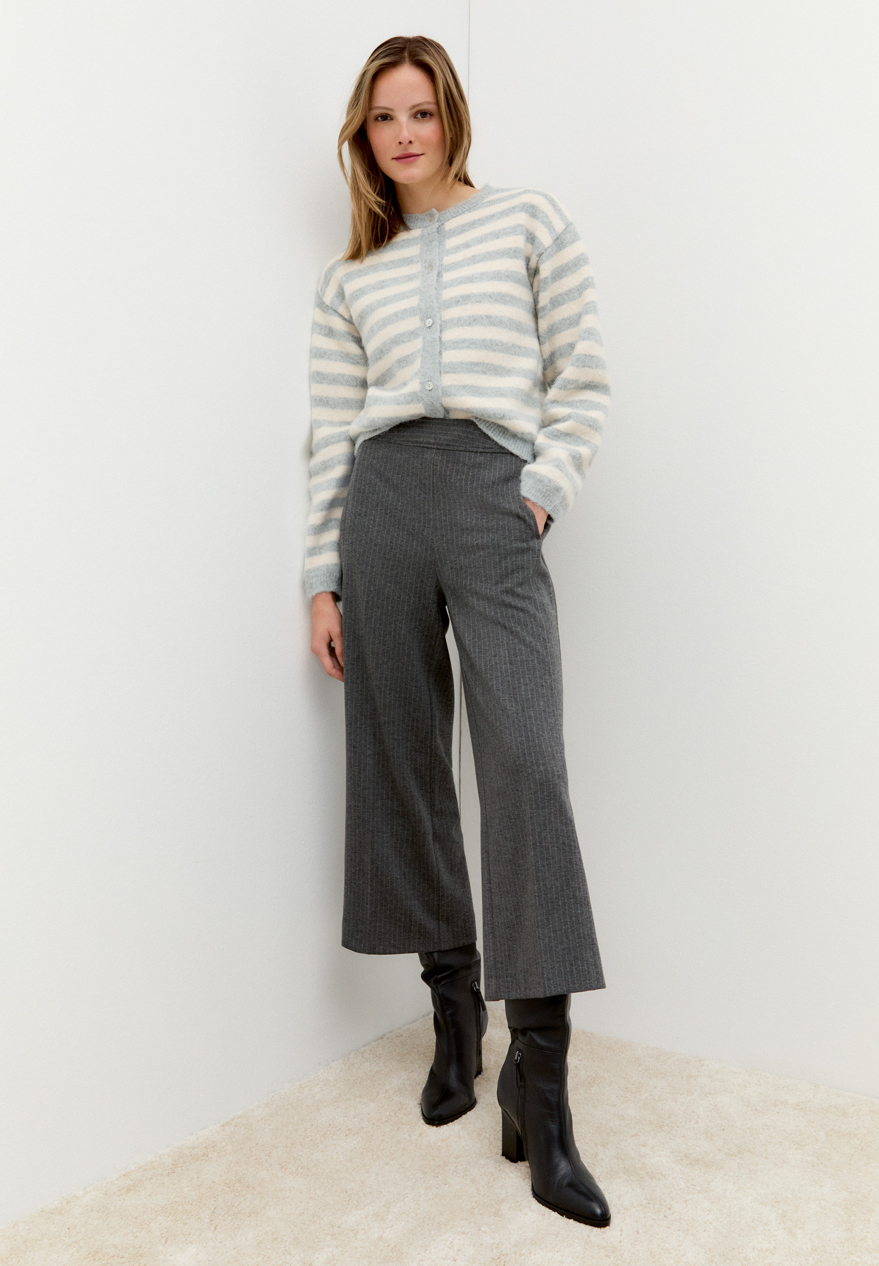 Eine Frau mit MORE & MORE Oberteil mit grauen Hosen und schwarzen Stiefeln vor einer weißen Wand. Eine Frau mit MORE & MORE Oberteil mit grauen Hosen und schwarzen Stiefeln vor einer weißen Wand.