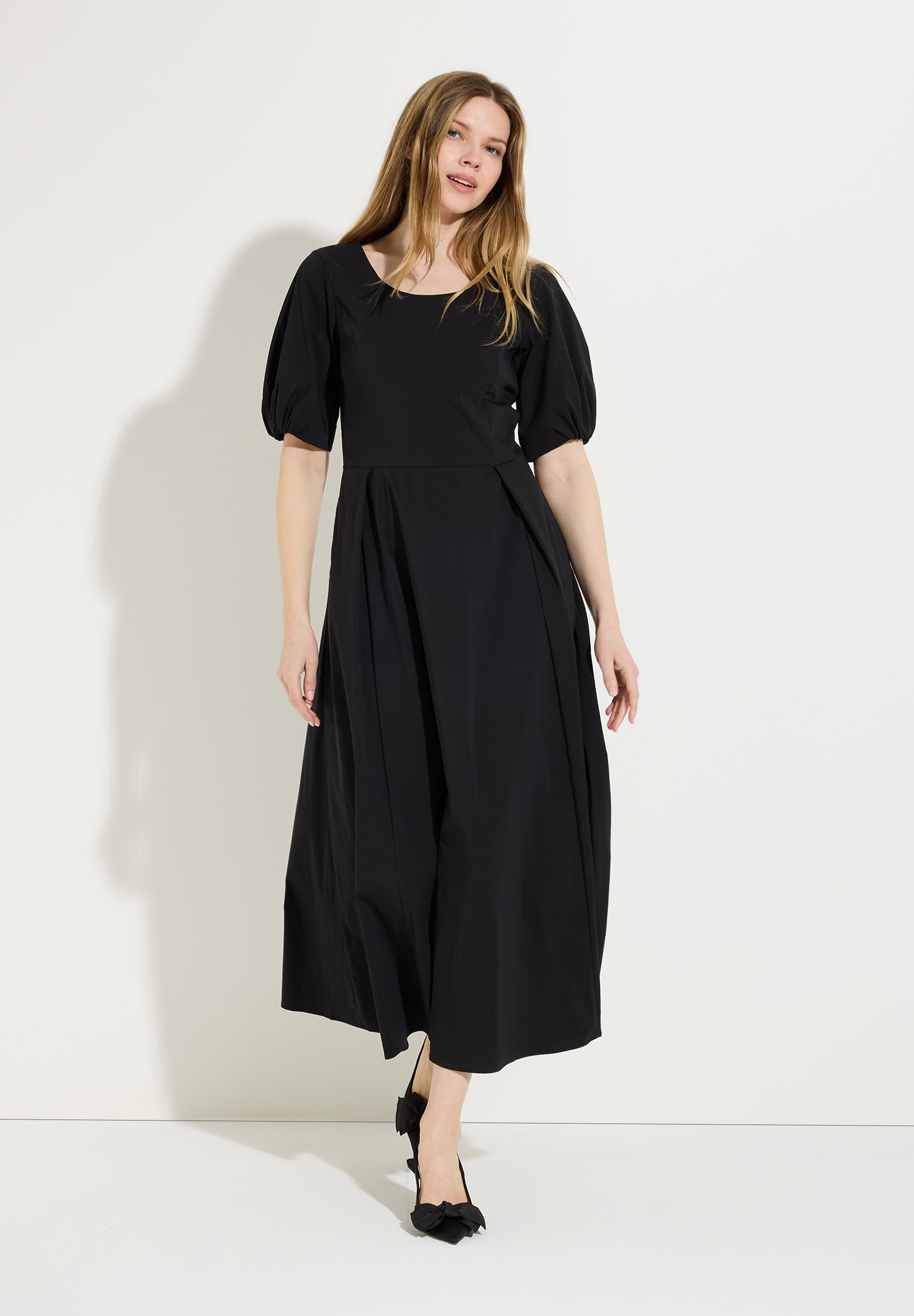 Eine Frau präsentiert ein schwarzes Kleid von MORE & MORE mit Popeline und Puffärmeln vor hellem Hintergrund.