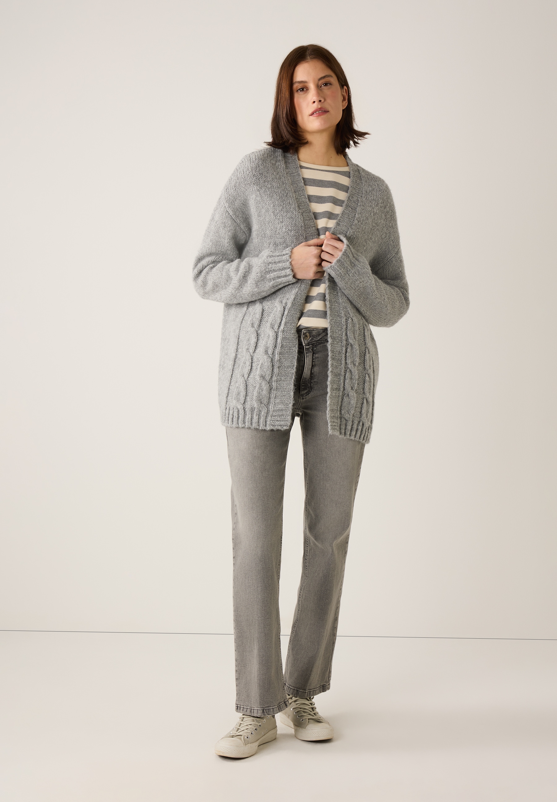 Eine Frau in grauer Strickjacke von MORE & MORE mit Zopfmuster, Jeans und Turnschuhen. Eine Frau in grauer Strickjacke von MORE & MORE mit Zopfmuster, Jeans und Turnschuhen.