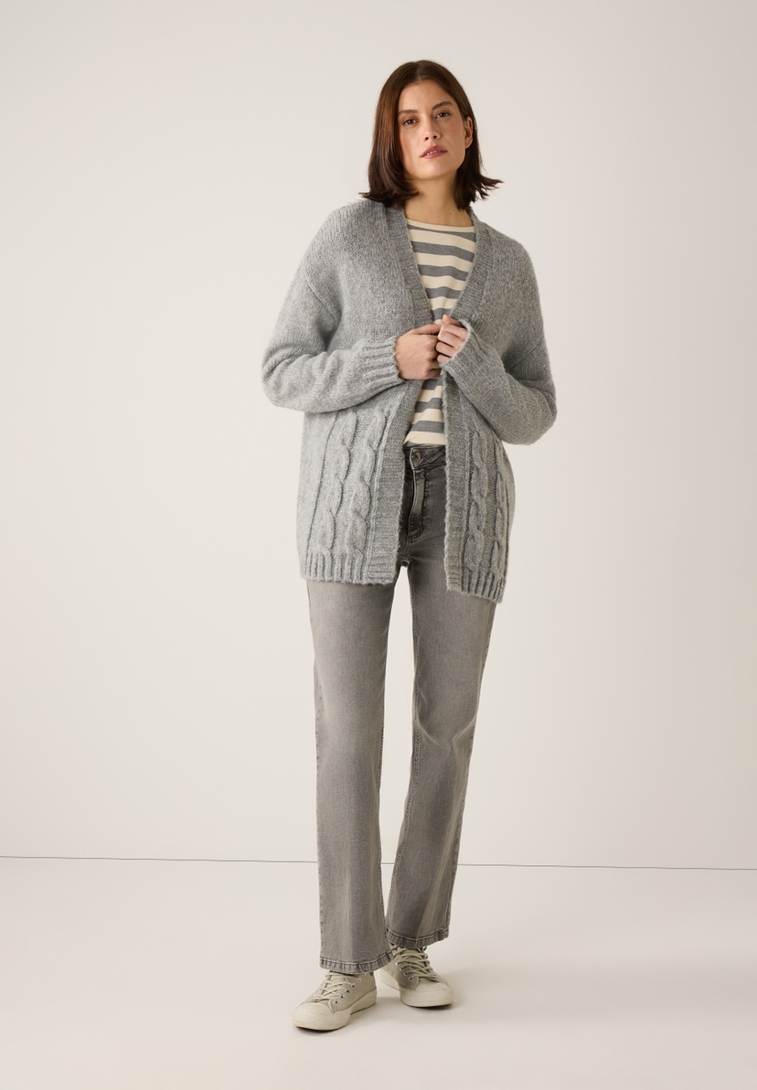 Eine Frau in grauer Strickjacke von MORE & MORE mit Zopfmuster, Jeans und Turnschuhen. Eine Frau in grauer Strickjacke von MORE & MORE mit Zopfmuster, Jeans und Turnschuhen.