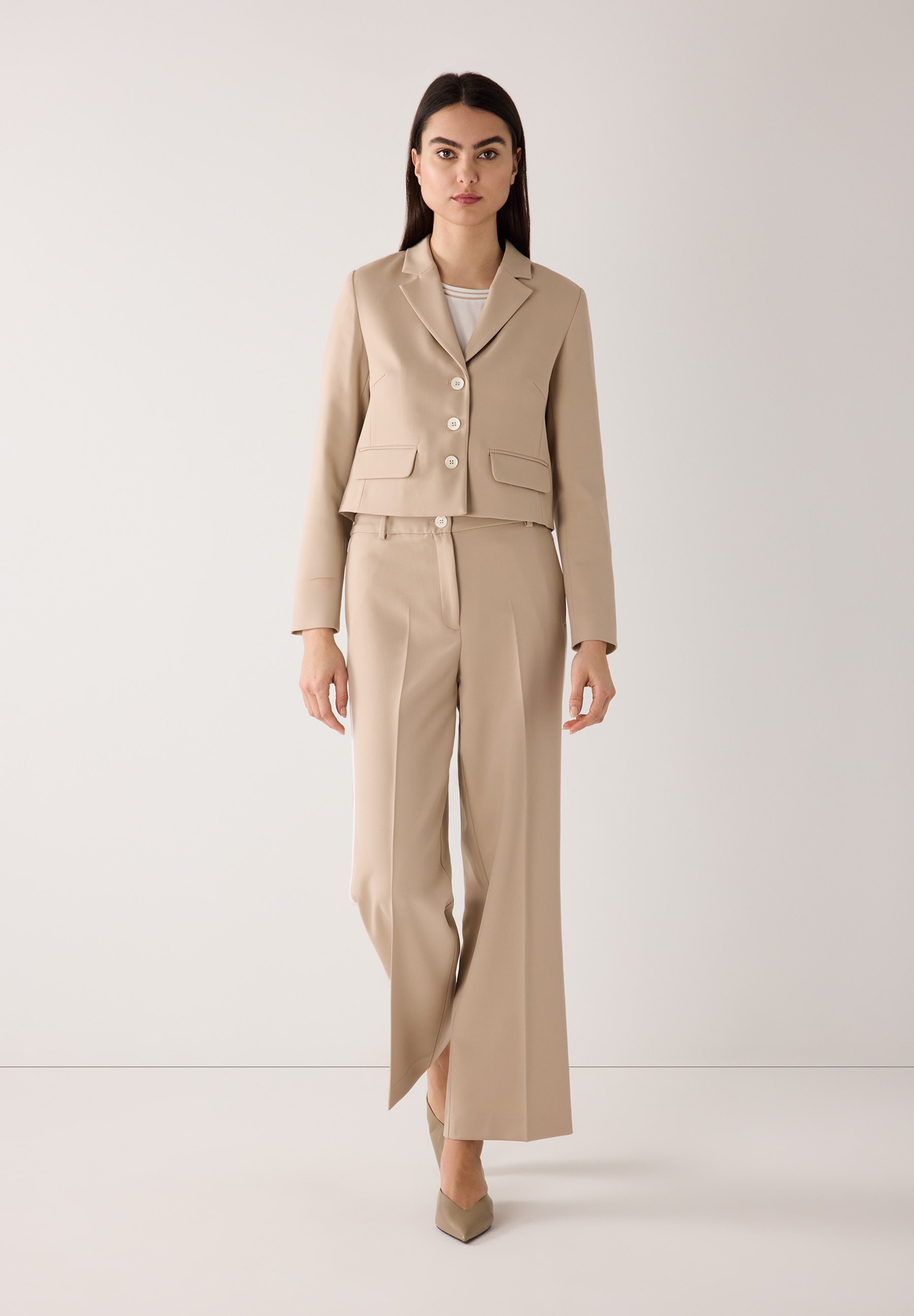 Eine Frau trägt einen beigen MORE & MORE Blazer mit passender Hose vor einem weißen Hintergrund. Eine Frau trägt einen beigen MORE & MORE Blazer mit passender Hose vor einem weißen Hintergrund.