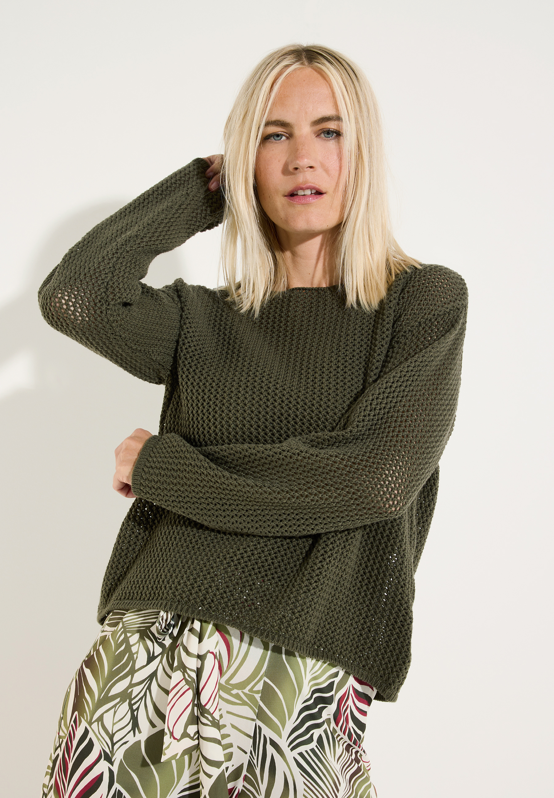 MORE & MORE Pullover: Eine blonde Frau trägt einen khakifarbenen, locker sitzenden Strickpullover und eine Hose mit Blättermuster. MORE & MORE Pullover: Eine blonde Frau trägt einen khakifarbenen, locker sitzenden Strickpullover und eine Hose mit Blättermuster.