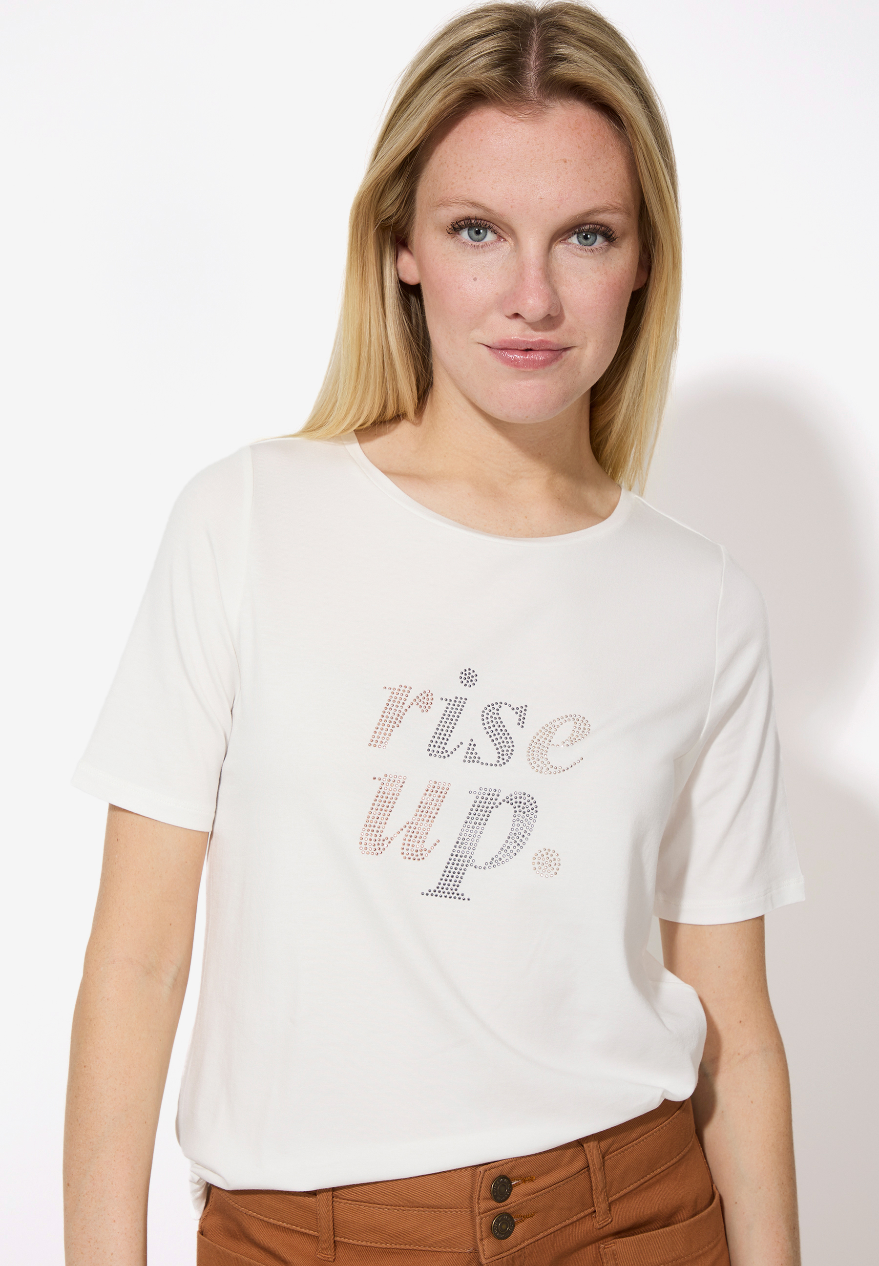 Eine Frau mit blonden Haaren trägt ein weißes T-Shirt von MORE & MORE mit dem Aufdruck "rise up".