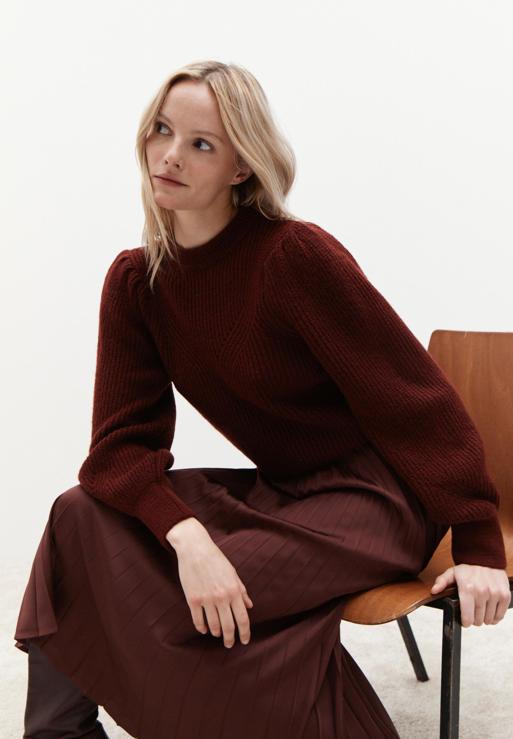 Eine Frau sitzt auf einem Stuhl und trägt einen bordeauxroten Pullover von MORE & MORE und einen passenden Rock. Eine Frau sitzt auf einem Stuhl und trägt einen bordeauxroten Pullover von MORE & MORE und einen passenden Rock.