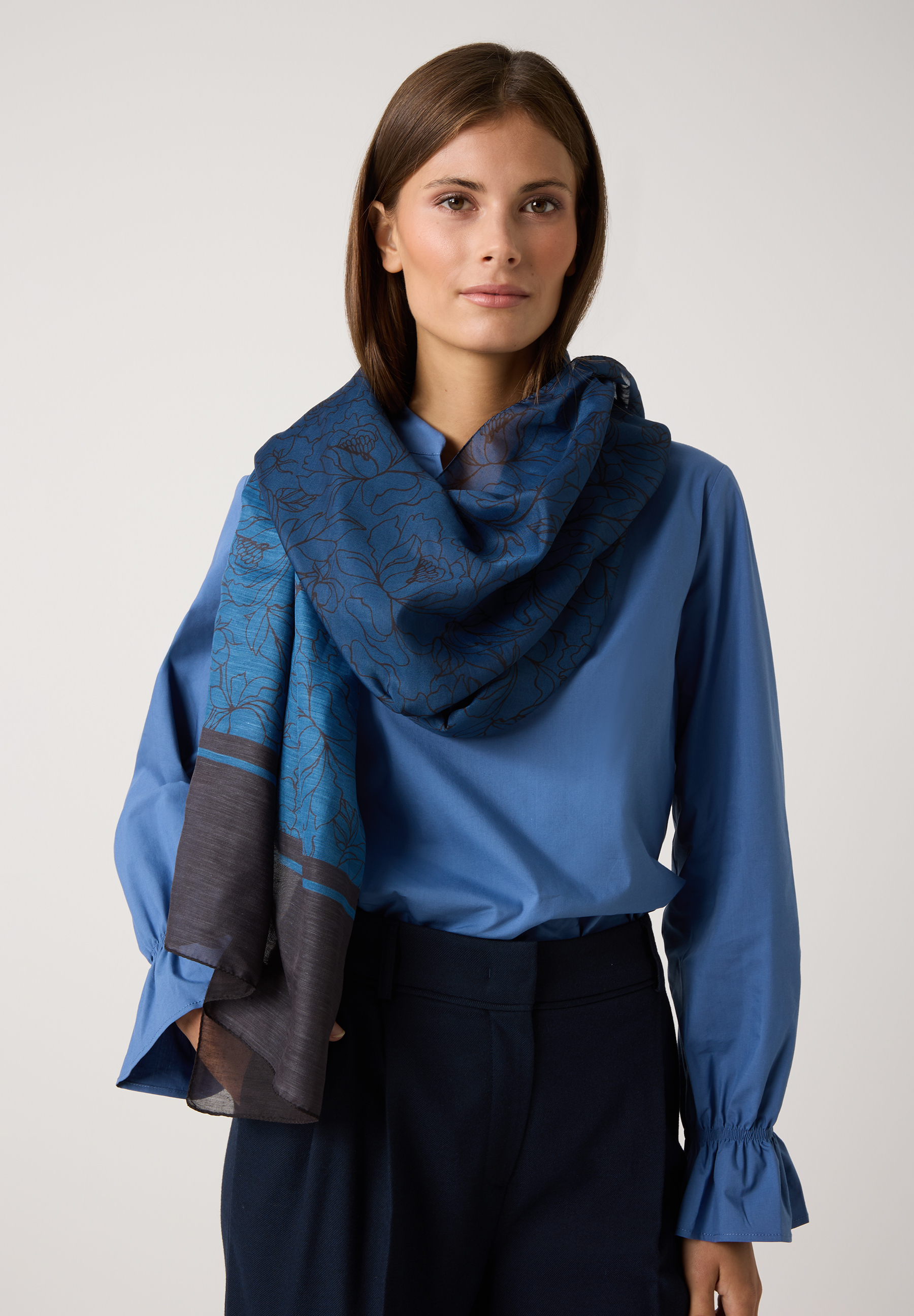 Eine Frau mit MORE & MORE Accessoire: blau gemusterter Schal zu blauer Bluse und dunkler Hose.