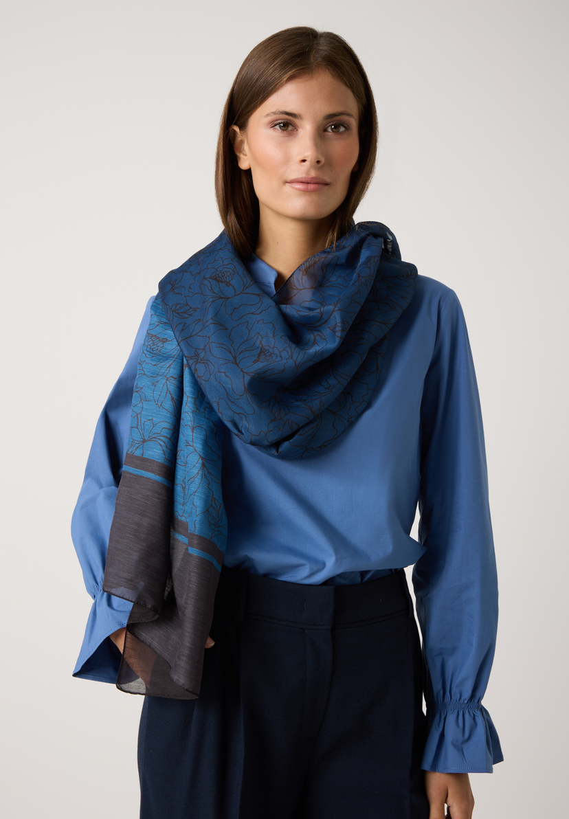 Eine Frau mit MORE & MORE Accessoire: blau gemusterter Schal zu blauer Bluse und dunkler Hose. Eine Frau mit MORE & MORE Accessoire: blau gemusterter Schal zu blauer Bluse und dunkler Hose.