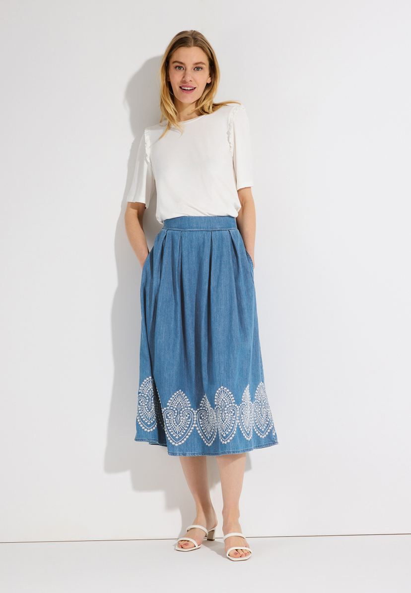 Eine Frau trägt einen blauen MORE & MORE Jeansrock mit weißem Muster am Saum. Eine Frau trägt einen blauen MORE & MORE Jeansrock mit weißem Muster am Saum.