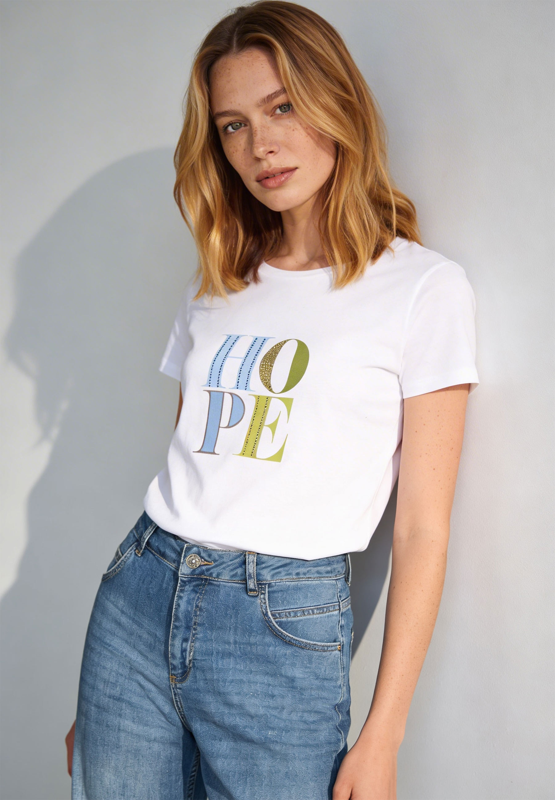 Eine Frau mit Sommersprossen trägt ein weißes MORE & MORE T-Shirt mit dem Aufdruck "HOPE" und Jeans.
