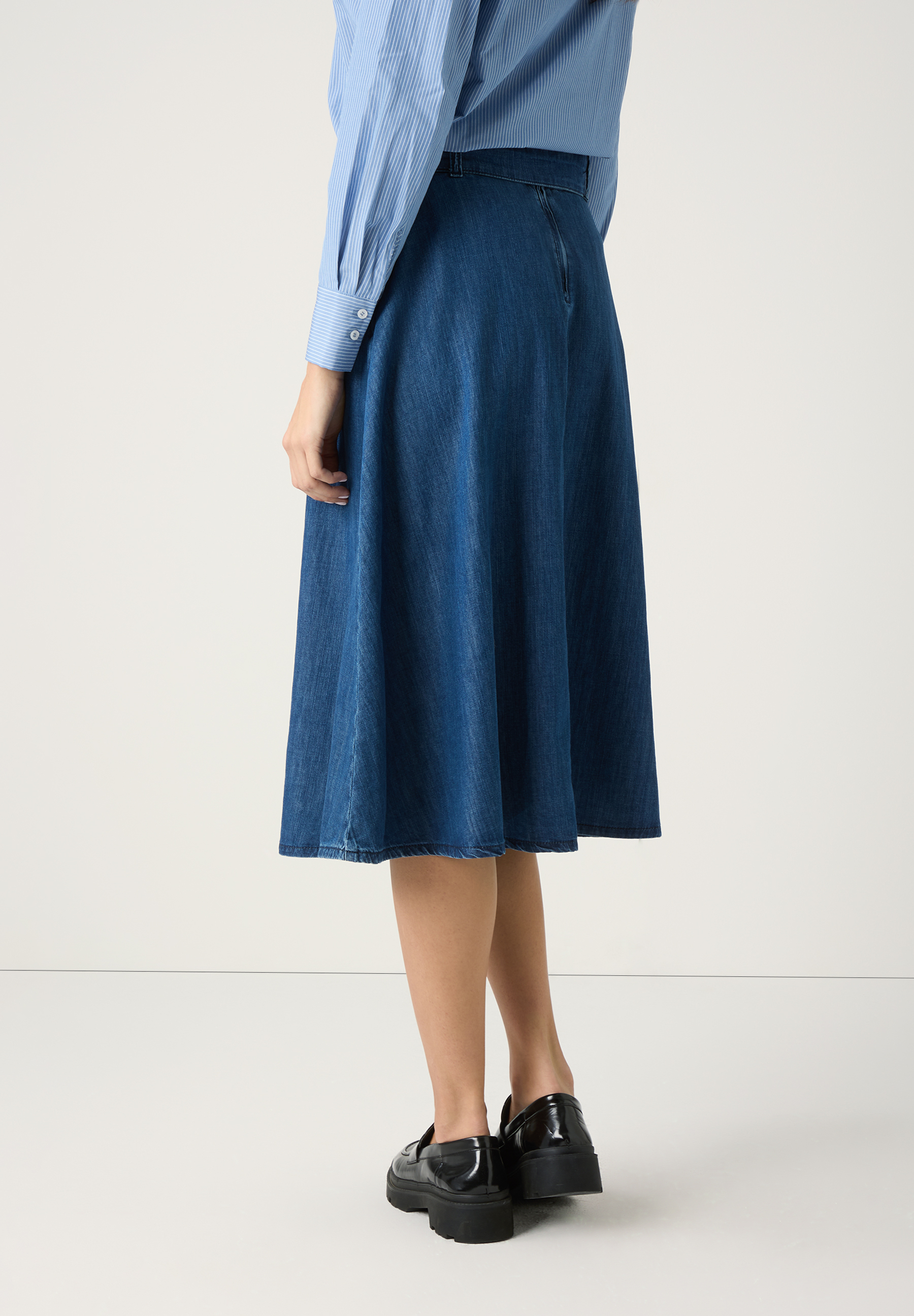 Ein MORE & MORE Rock aus Denim, kombiniert mit einem blau-weiß gestreiften Oberteil und schwarzen Schuhen, abgebildet von hinten. Ein MORE & MORE Rock aus Denim, kombiniert mit einem blau-weiß gestreiften Oberteil und schwarzen Schuhen, abgebildet von hinten.