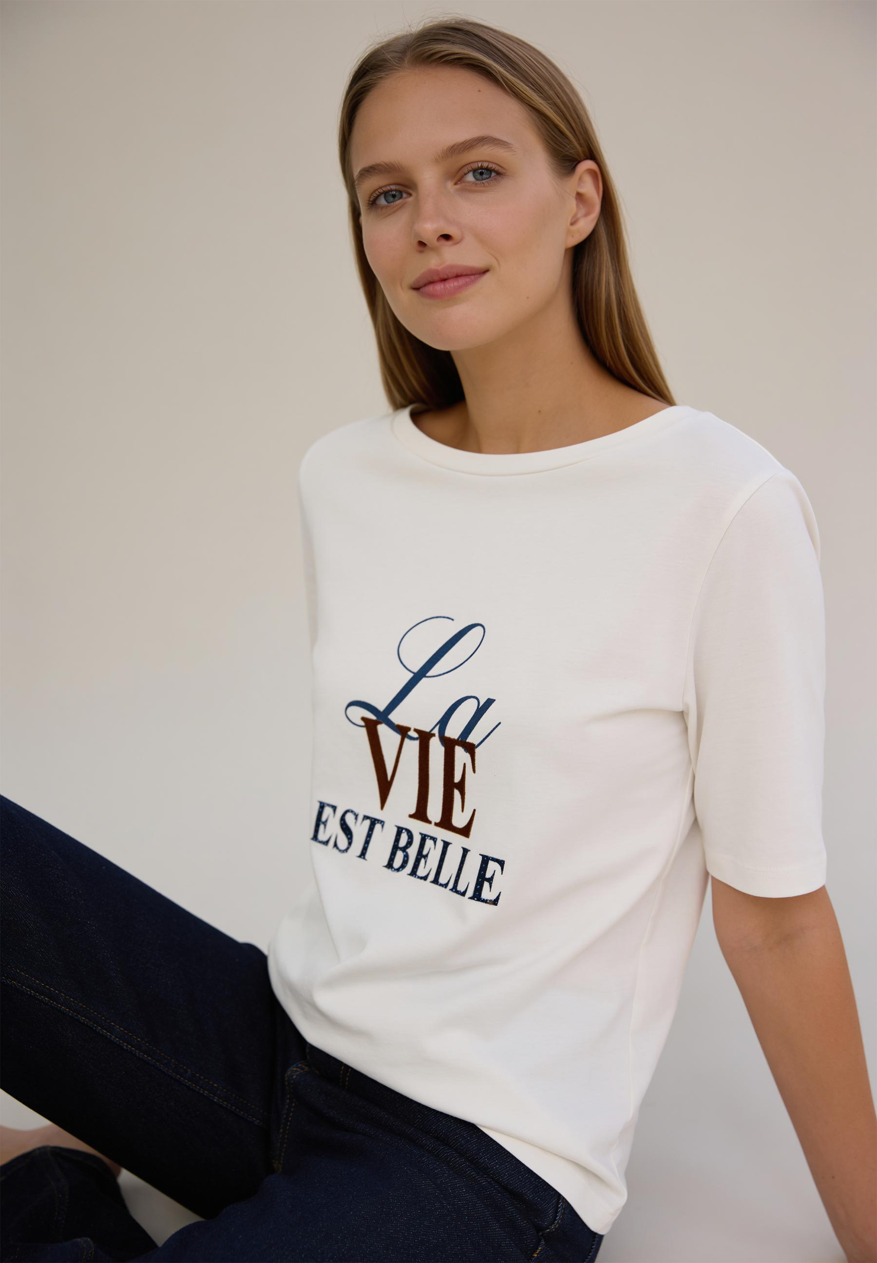 AI generated - MORE & MORE Damenshirt aus Jersey, cremefarben, mit Aufschrift "La vie est belle" in dunkelblauen/braunen Buchstaben.