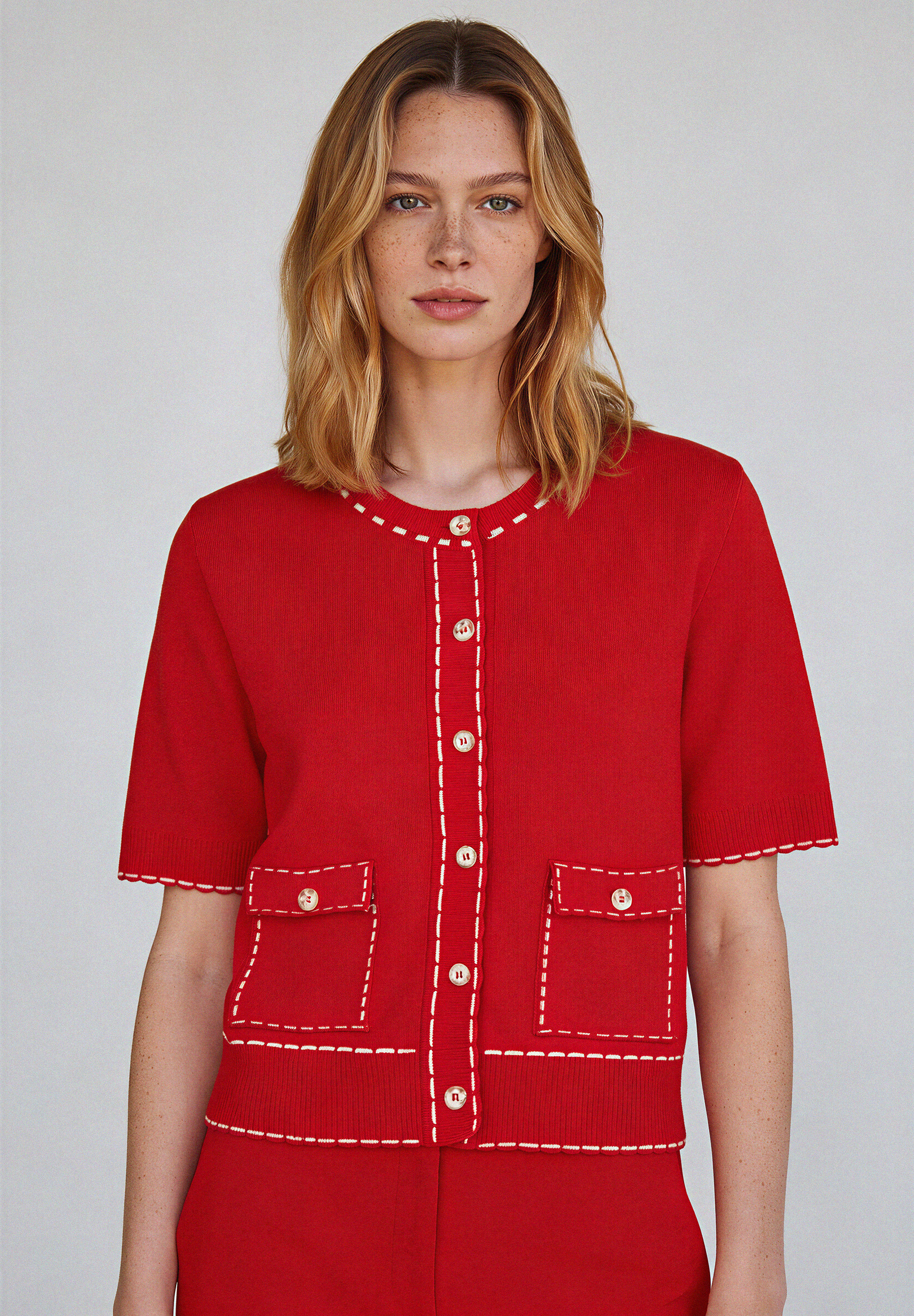 Eine Frau trägt einen roten Strick-Cardigan von MORE & MORE mit kurzen Ärmeln und aufgesetzten Taschen.