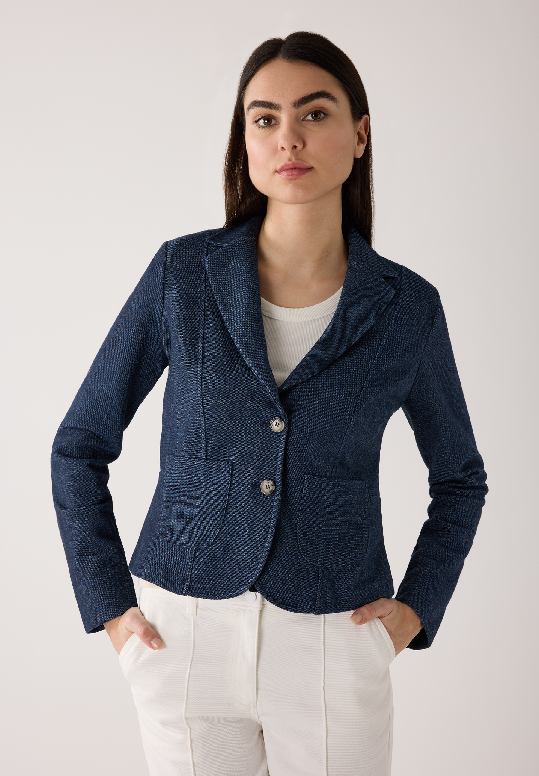 Eine Frau mit MORE & MORE Blazer und weißen Hosen posiert vor einem hellen Hintergrund.