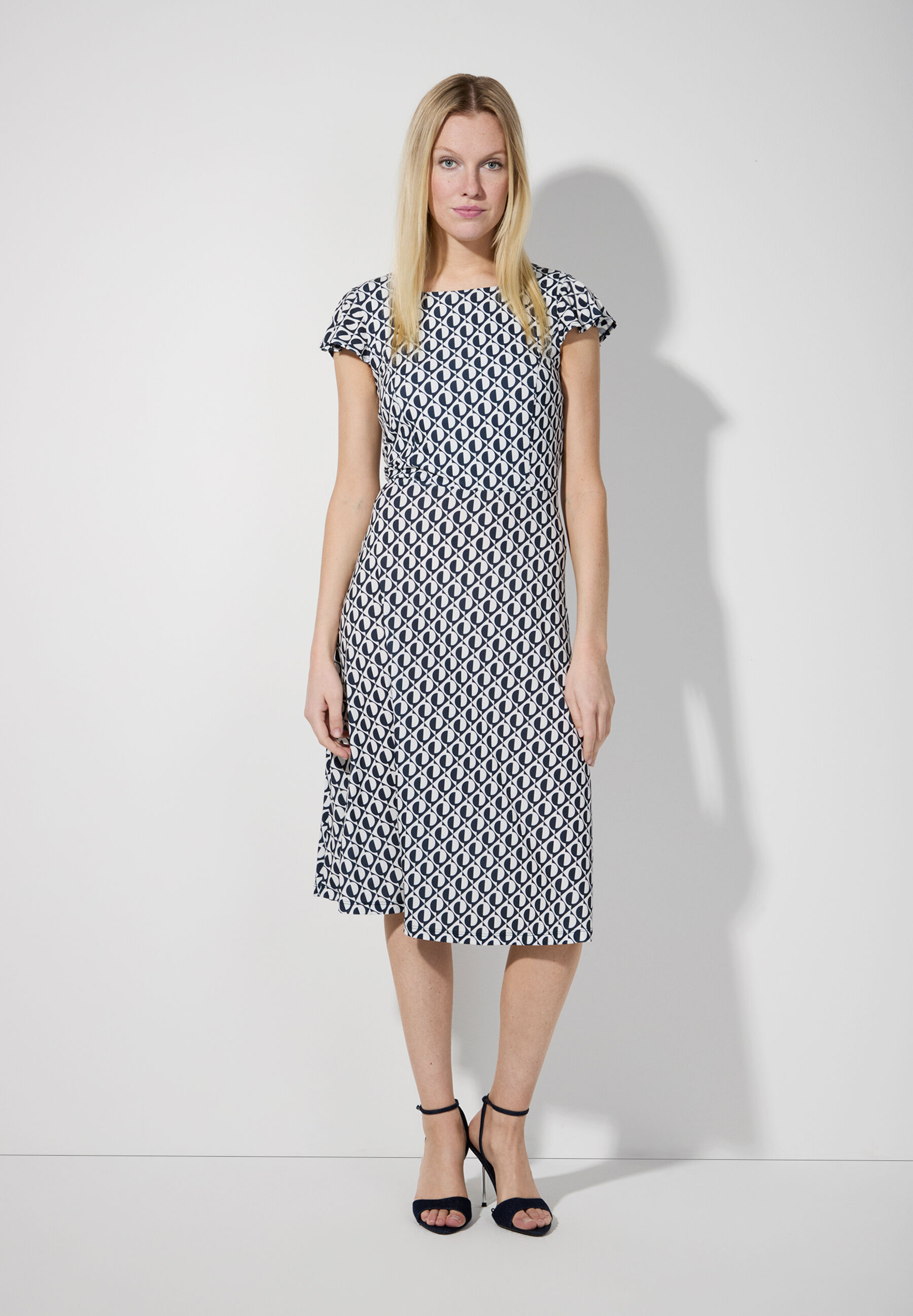 Eine blonde Frau trägt ein MORE & MORE Jersey-Kleid mit geometrischem Muster und Absatzschuhe. Eine blonde Frau trägt ein MORE & MORE Jersey-Kleid mit geometrischem Muster und Absatzschuhe.