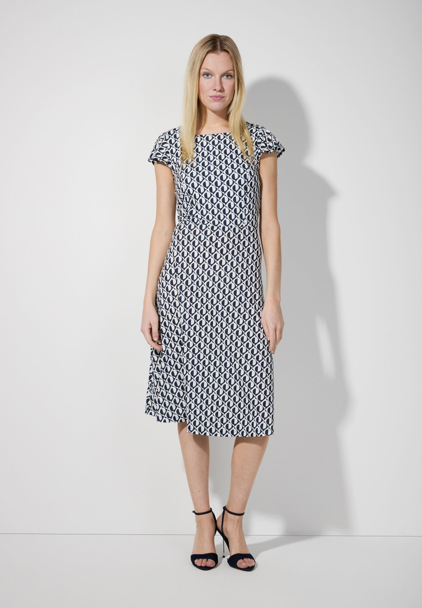 Eine blonde Frau trägt ein MORE & MORE Jersey-Kleid mit geometrischem Muster und Absatzschuhe. Eine blonde Frau trägt ein MORE & MORE Jersey-Kleid mit geometrischem Muster und Absatzschuhe.