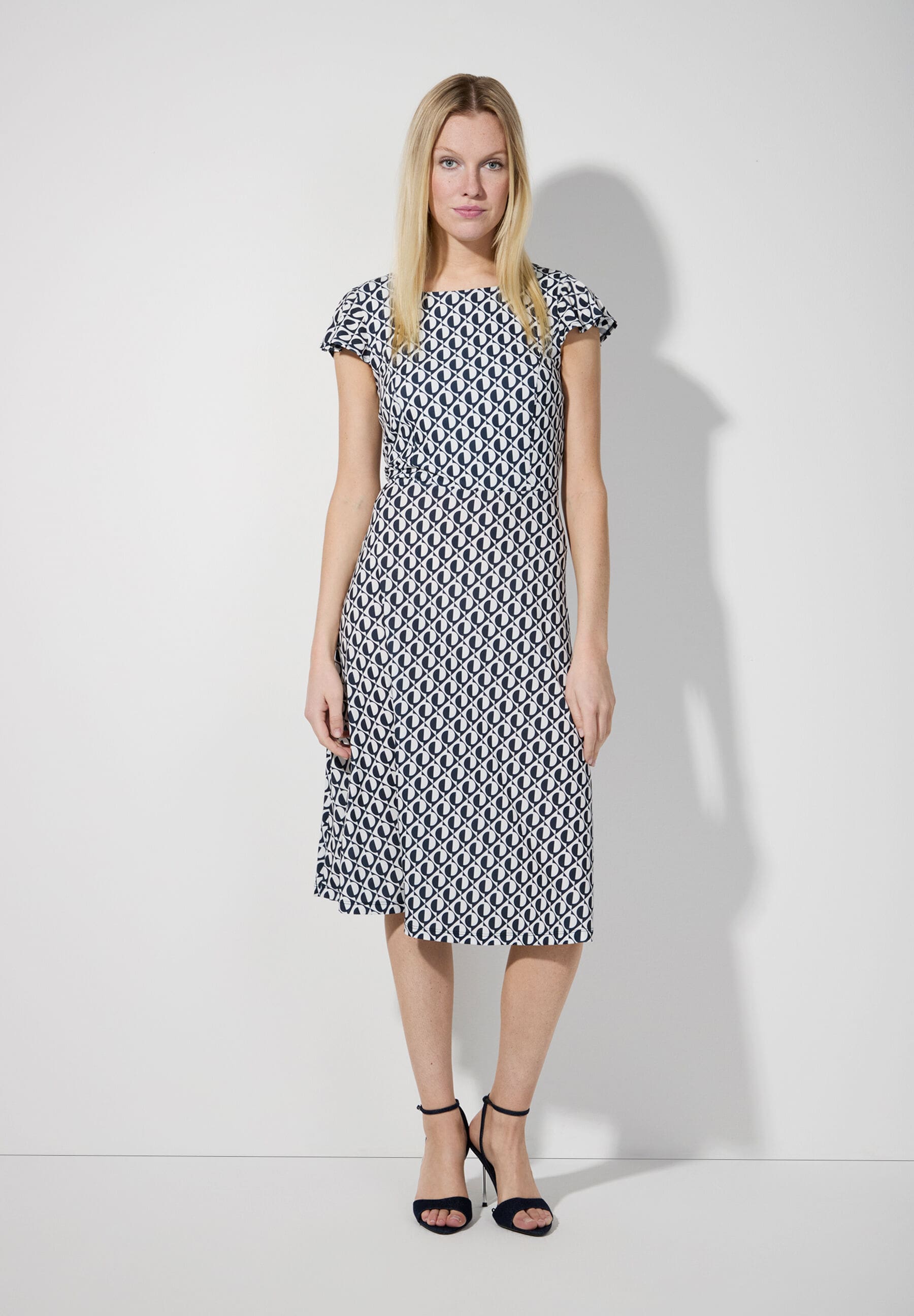 Eine blonde Frau trägt ein MORE & MORE Jersey-Kleid mit geometrischem Muster und Absatzschuhe.