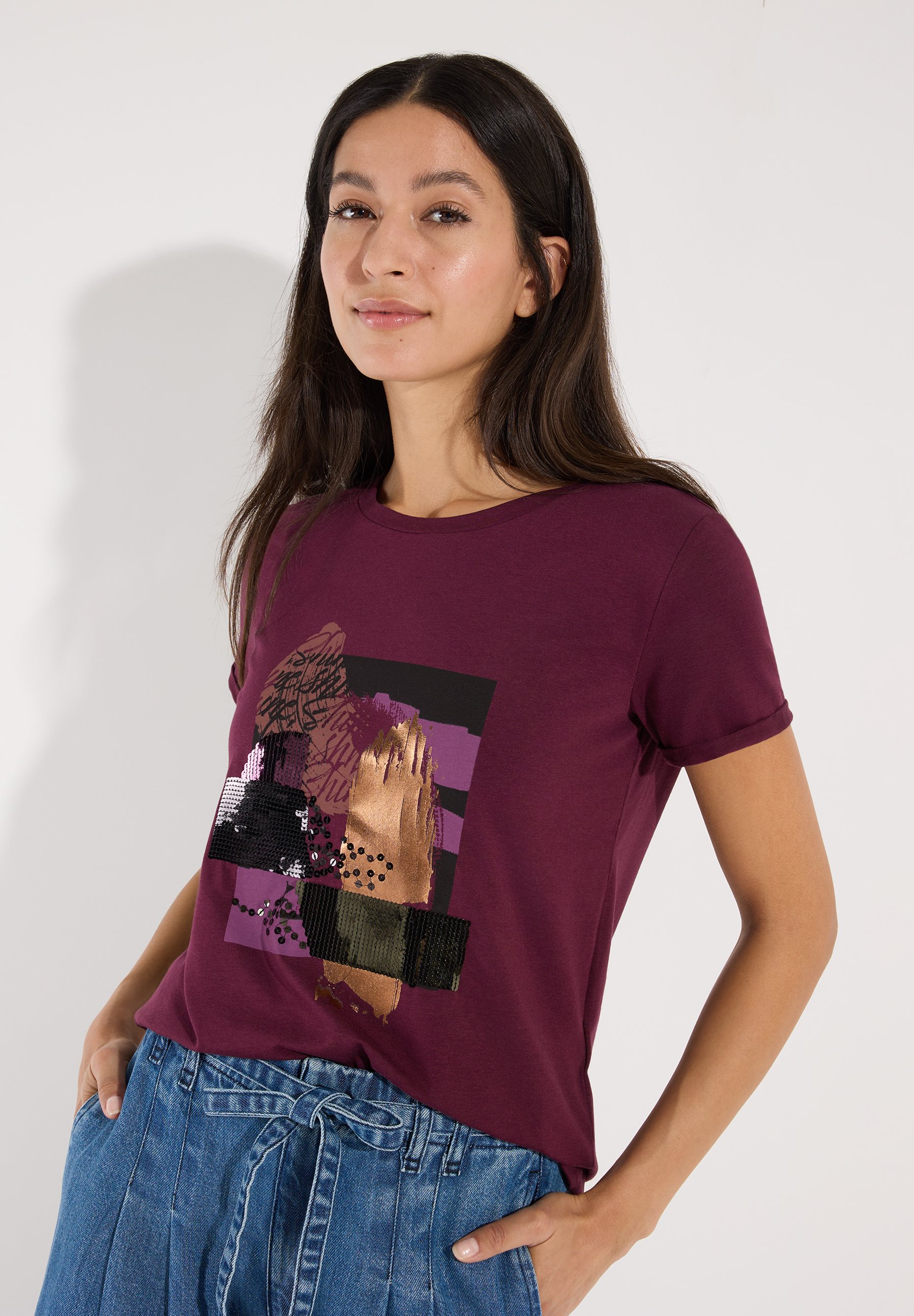 MORE & MORE T-Shirt in Dunkelrot mit glitzerndem, abstraktem Druck und Pailletten, kombiniert mit blauer Jeans.