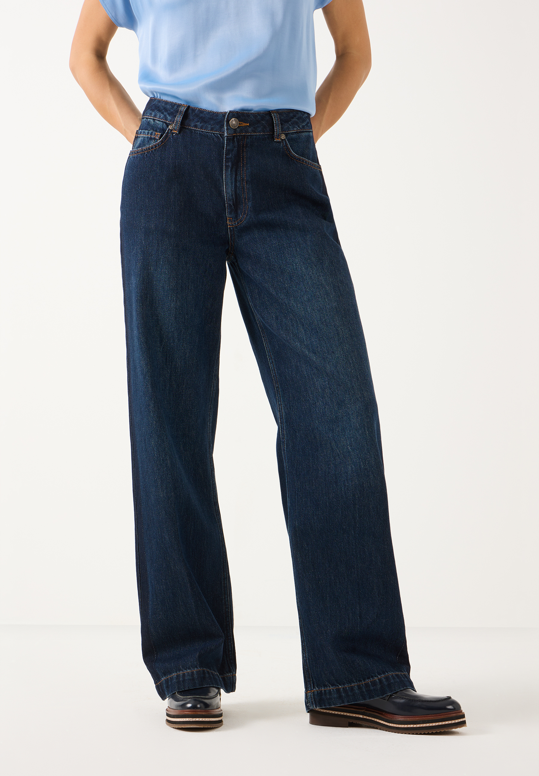 MORE & MORE Jeans: eine Person in dunkelblauer Denim-Jeans mit weitem Bein und hellem Oberteil, Hände in Hosentaschen.