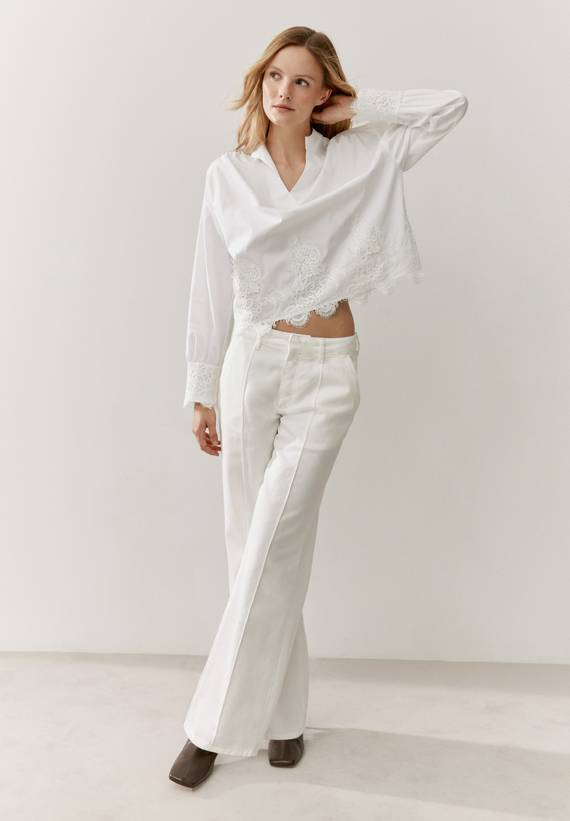Eine Frau trägt eine weiße MORE & MORE Bluse mit Spitze und eine weiße Hose vor einem hellen Hintergrund. Eine Frau trägt eine weiße MORE & MORE Bluse mit Spitze und eine weiße Hose vor einem hellen Hintergrund.