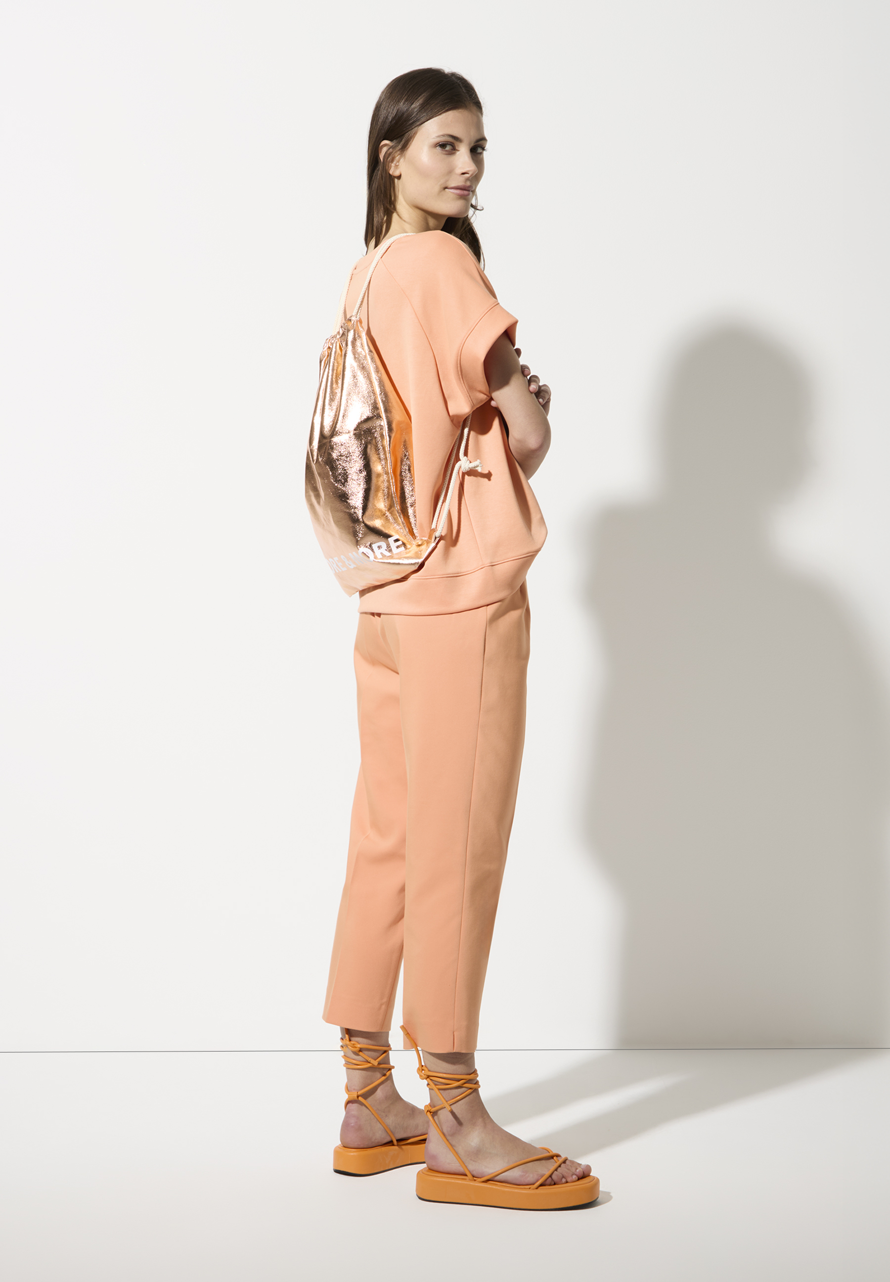 Eine Frau mit MORE & MORE Sweatshirt und Hose in Pfirsichfarbe, dazu goldener Rucksack. Eine Frau mit MORE & MORE Sweatshirt und Hose in Pfirsichfarbe, dazu goldener Rucksack.