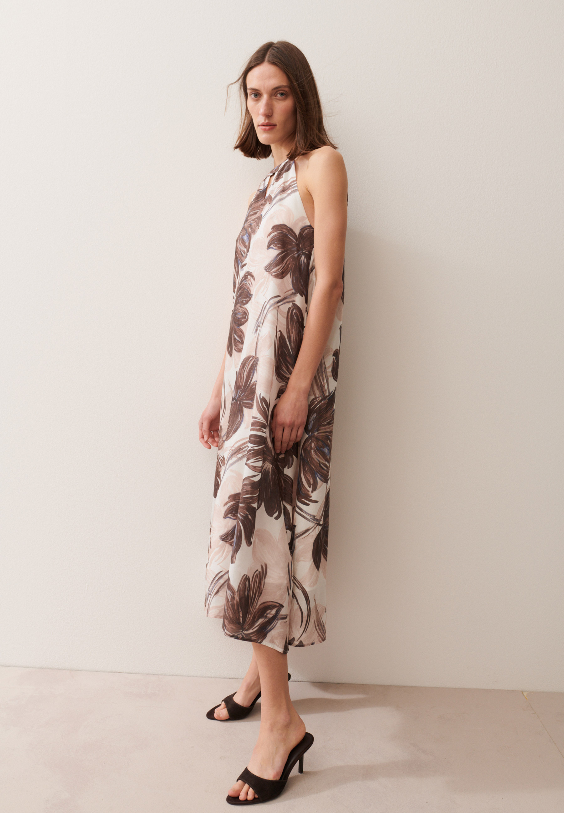 Eine Frau zeigt ein ärmelloses MORE & MORE Kleid mit braunen Blumen und dazu schwarze High Heels.