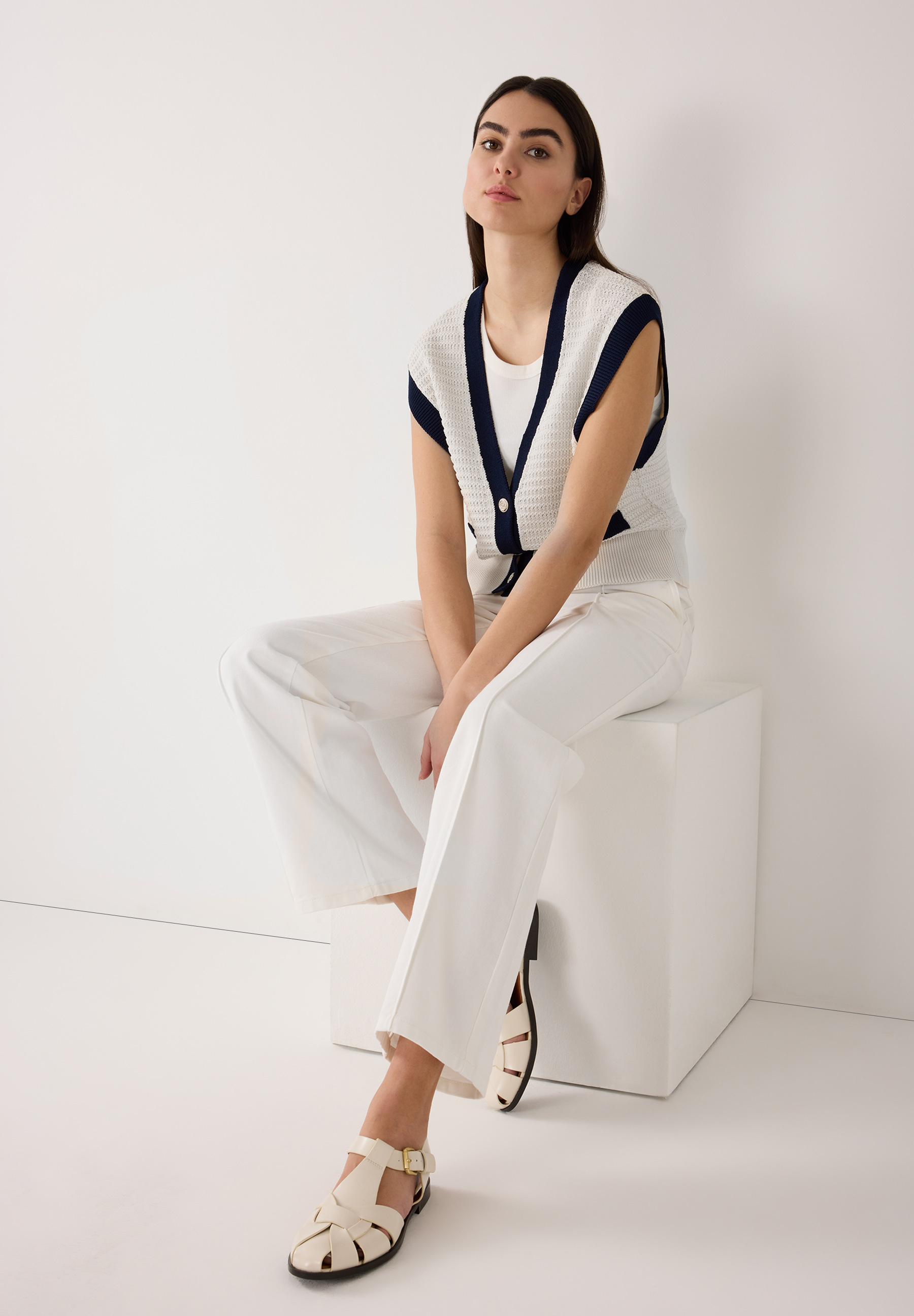 Eine Frau in weißer MORE & MORE Strickmode sitzt auf einem weißen Hocker vor weißem Hintergrund. Eine Frau in weißer MORE & MORE Strickmode sitzt auf einem weißen Hocker vor weißem Hintergrund.