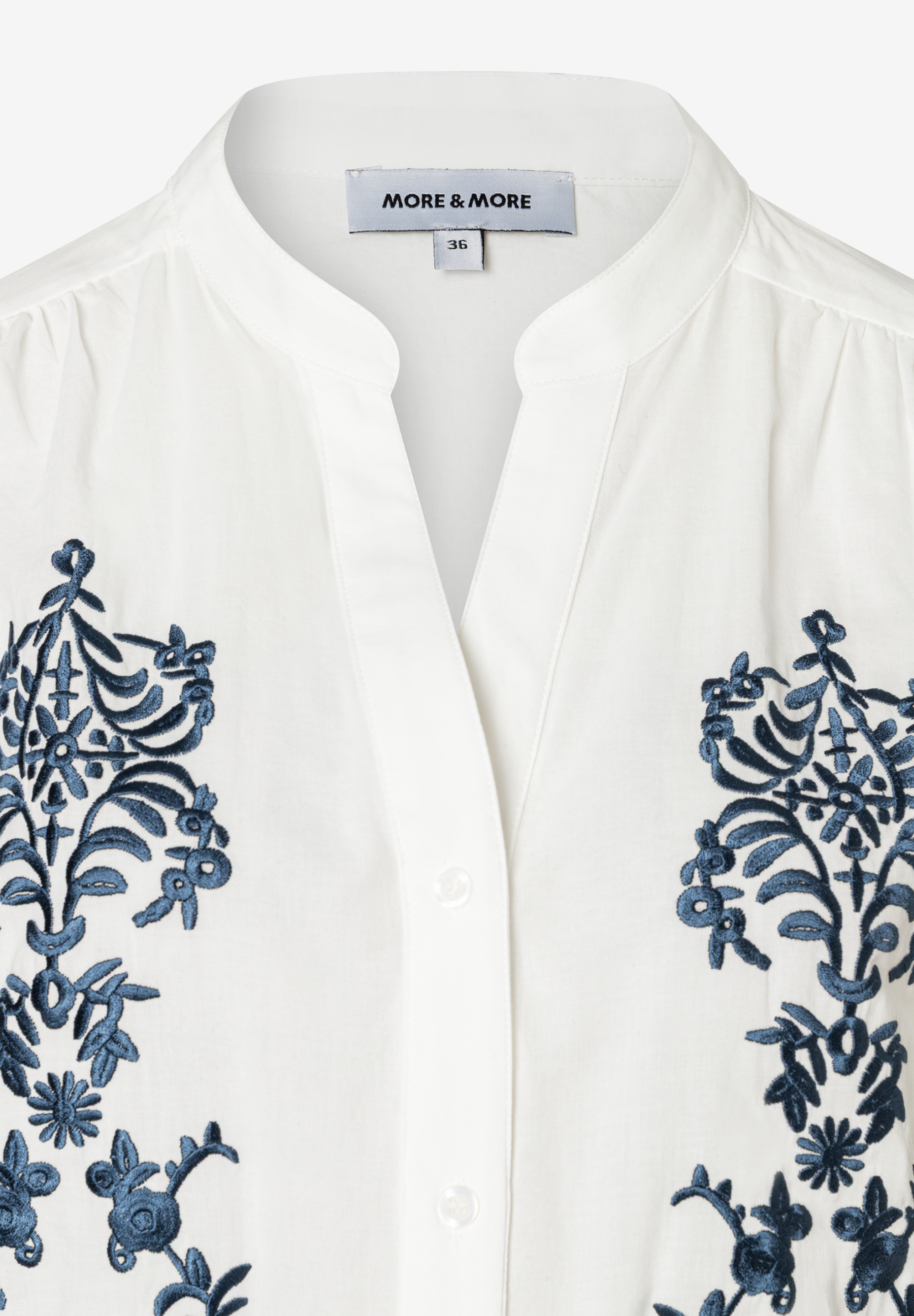 Detailansicht einer weißen Bluse von MORE & MORE mit blauen Stickereien und Knopfleiste. Detailansicht einer weißen Bluse von MORE & MORE mit blauen Stickereien und Knopfleiste.