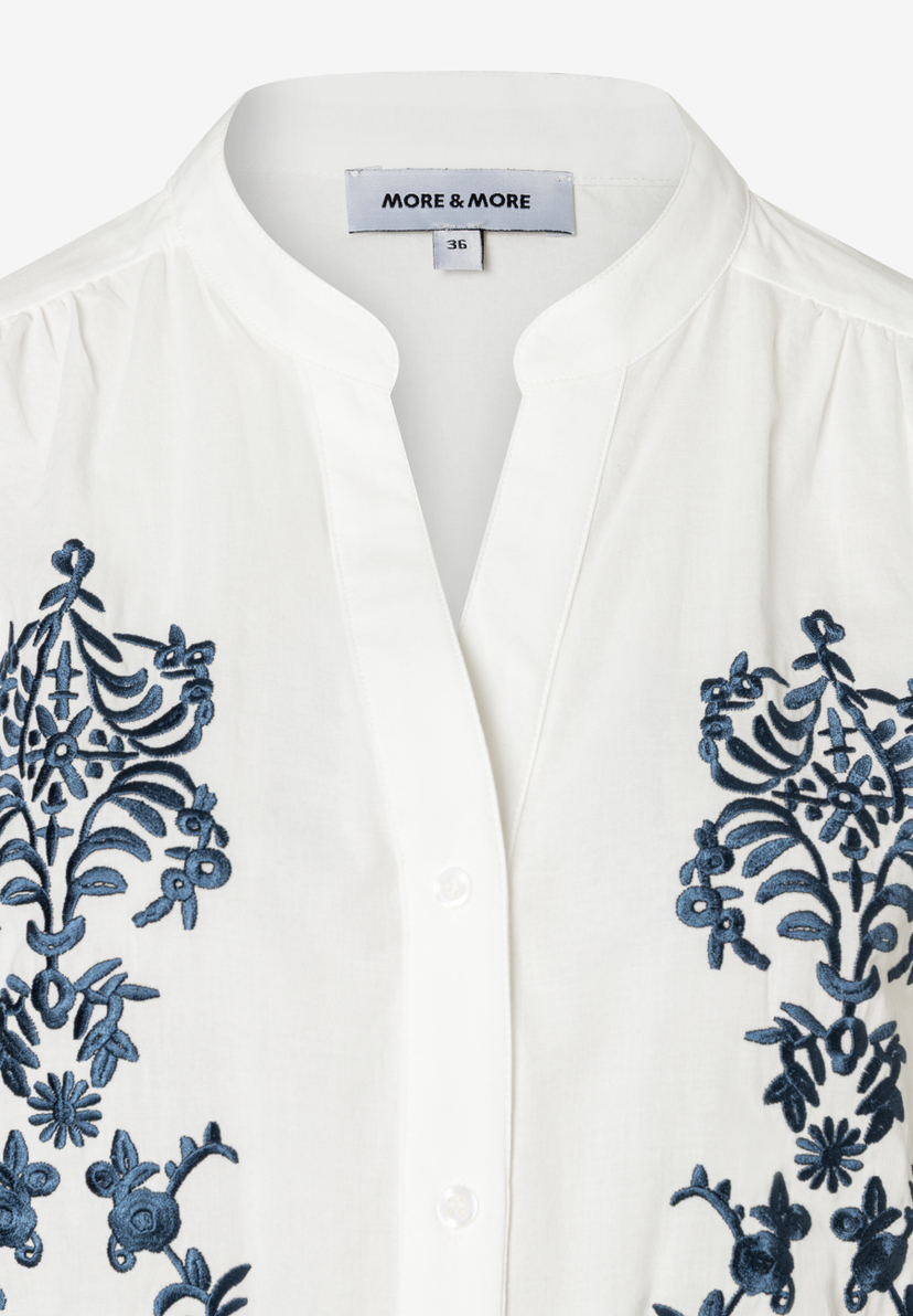 Detailansicht einer weißen Bluse von MORE & MORE mit blauen Stickereien und Knopfleiste. Detailansicht einer weißen Bluse von MORE & MORE mit blauen Stickereien und Knopfleiste.