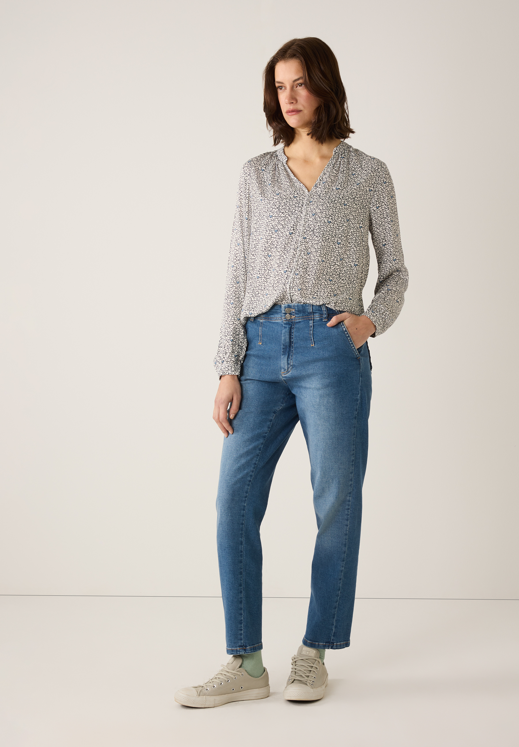 Eine Frau in einer MORE & MORE Bluse mit Herzmuster und Jeans steht vor einem hellen Hintergrund. Eine Frau in einer MORE & MORE Bluse mit Herzmuster und Jeans steht vor einem hellen Hintergrund.