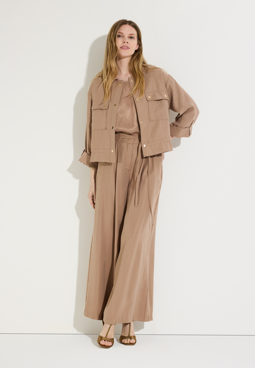 Eine Frau in MORE & MORE-Hosen steht vor einer weißen Wand. Sie trägt eine passende Jacke und Sandalen.