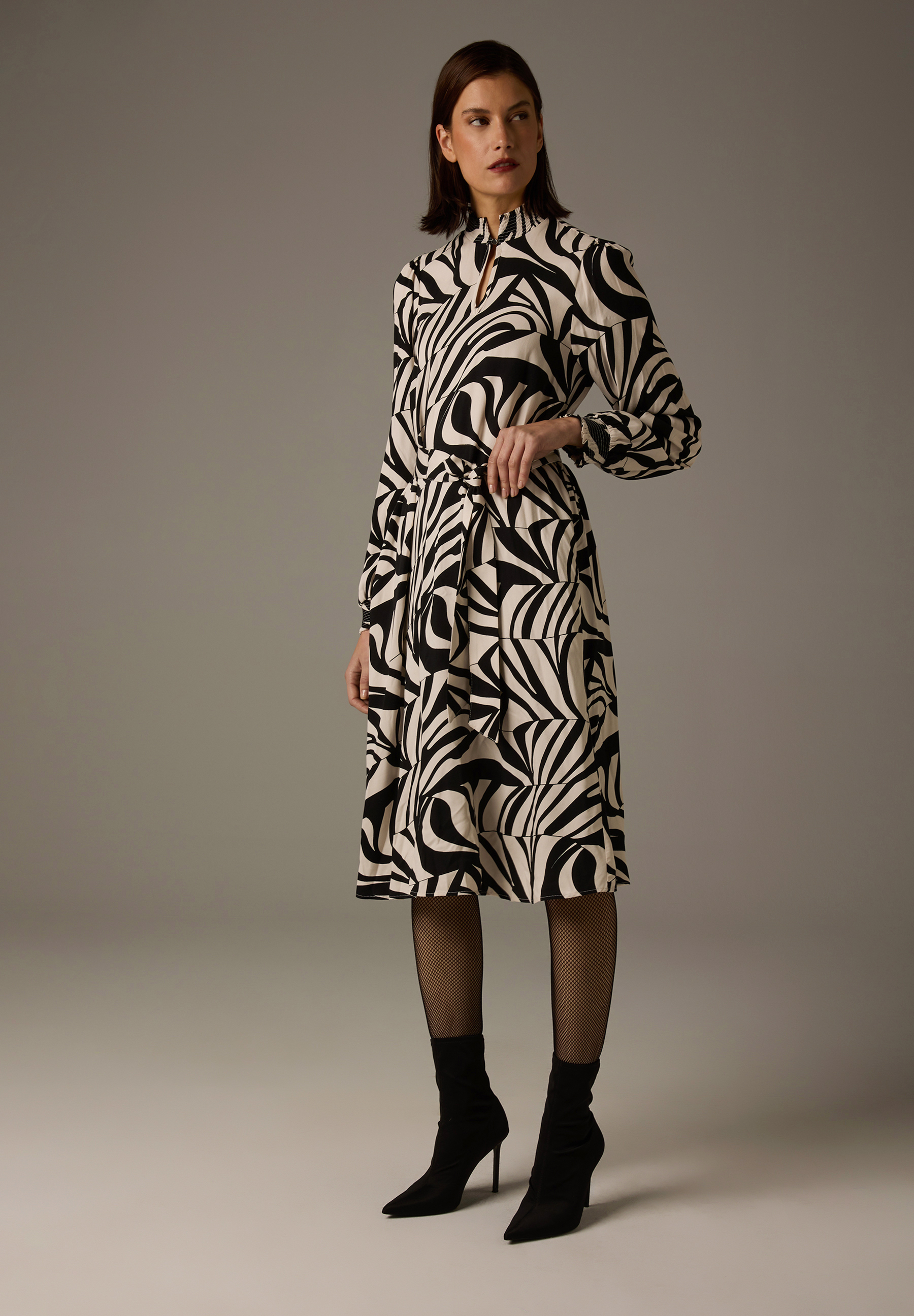 Eine Frau im MORE & MORE-Viskosekleid mit auffälligem Zebra-Muster, blickdichten Strumpfhosen und Stiefeln.