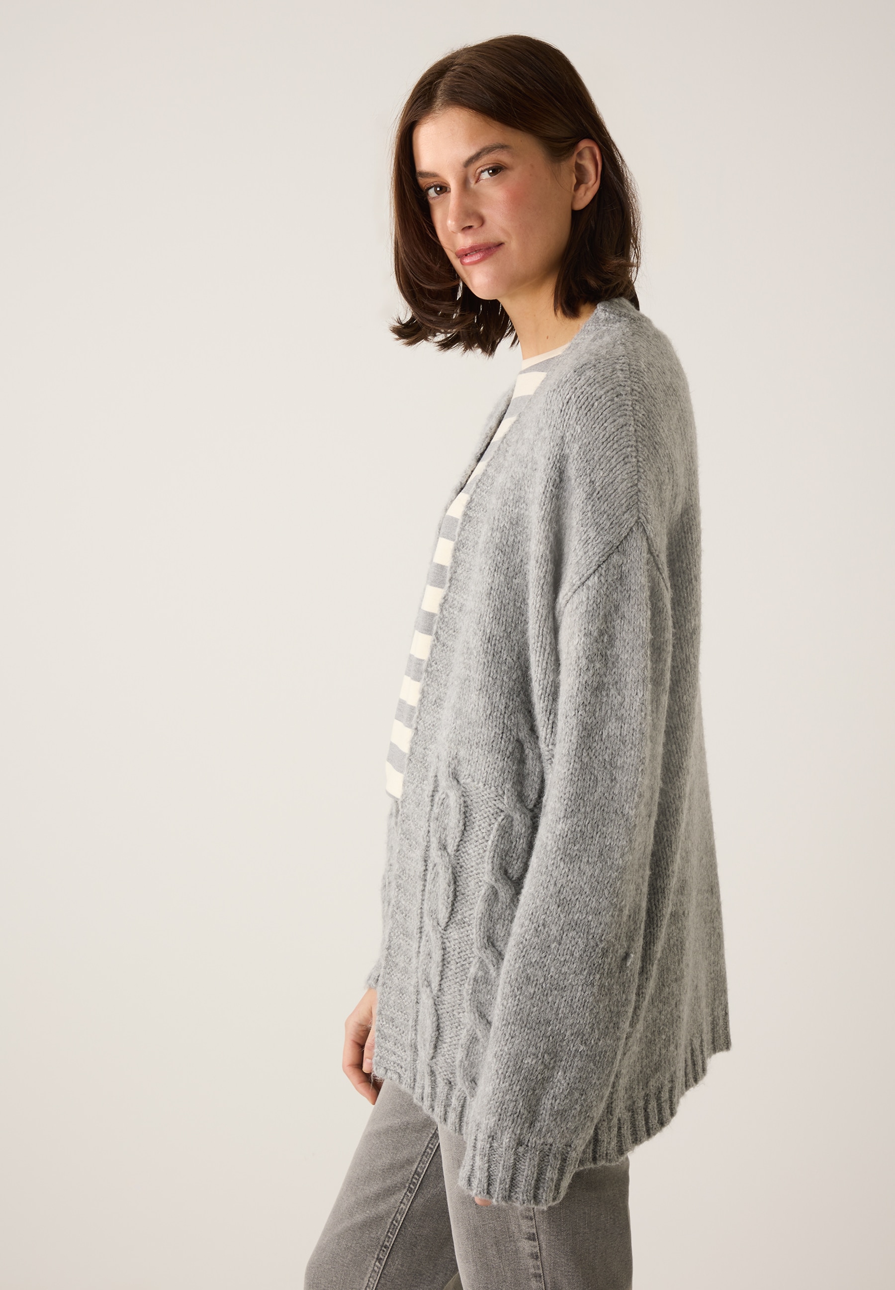 Eine Frau trägt eine graue Strickjacke der Marke MORE & MORE. Die Jacke hat ein Zopfmuster und wirkt angenehm wärmend. Eine Frau trägt eine graue Strickjacke der Marke MORE & MORE. Die Jacke hat ein Zopfmuster und wirkt angenehm wärmend.