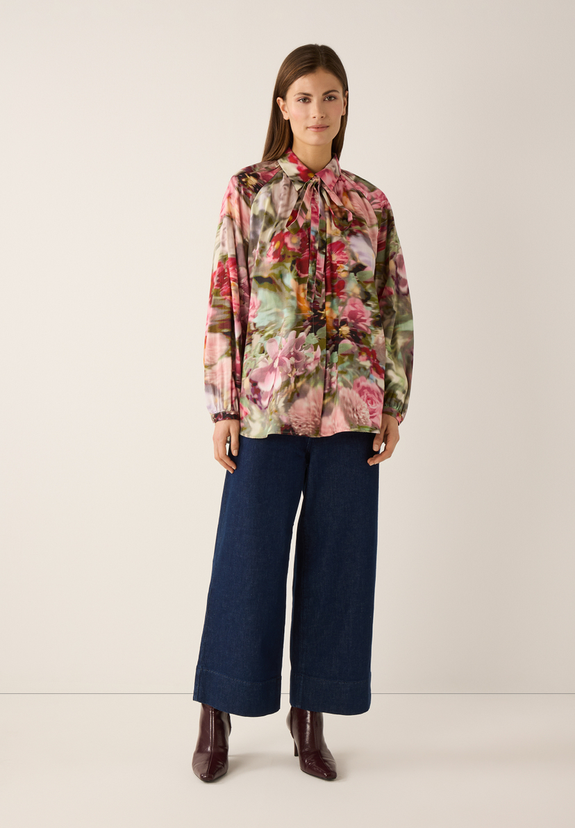 Eine Frau in MORE & MORE Jeans und floraler Bluse, posiert vor hellem Hintergrund. Eine Frau in MORE & MORE Jeans und floraler Bluse, posiert vor hellem Hintergrund.