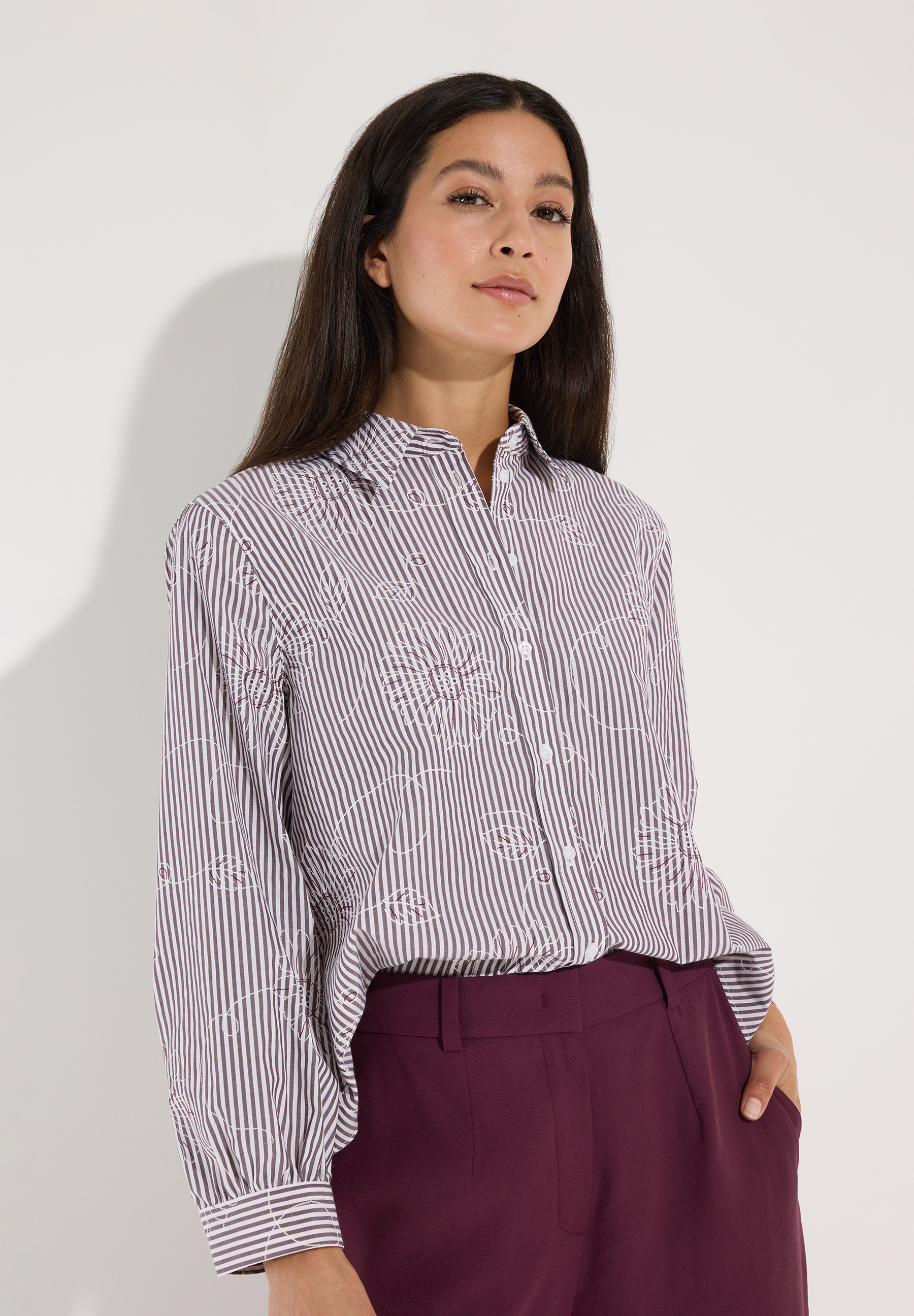 MORE & MORE Bluse: Eine Frau trägt eine gestreifte MORE & MORE Bluse mit floralen Stickereien und einer weinroten Hose.