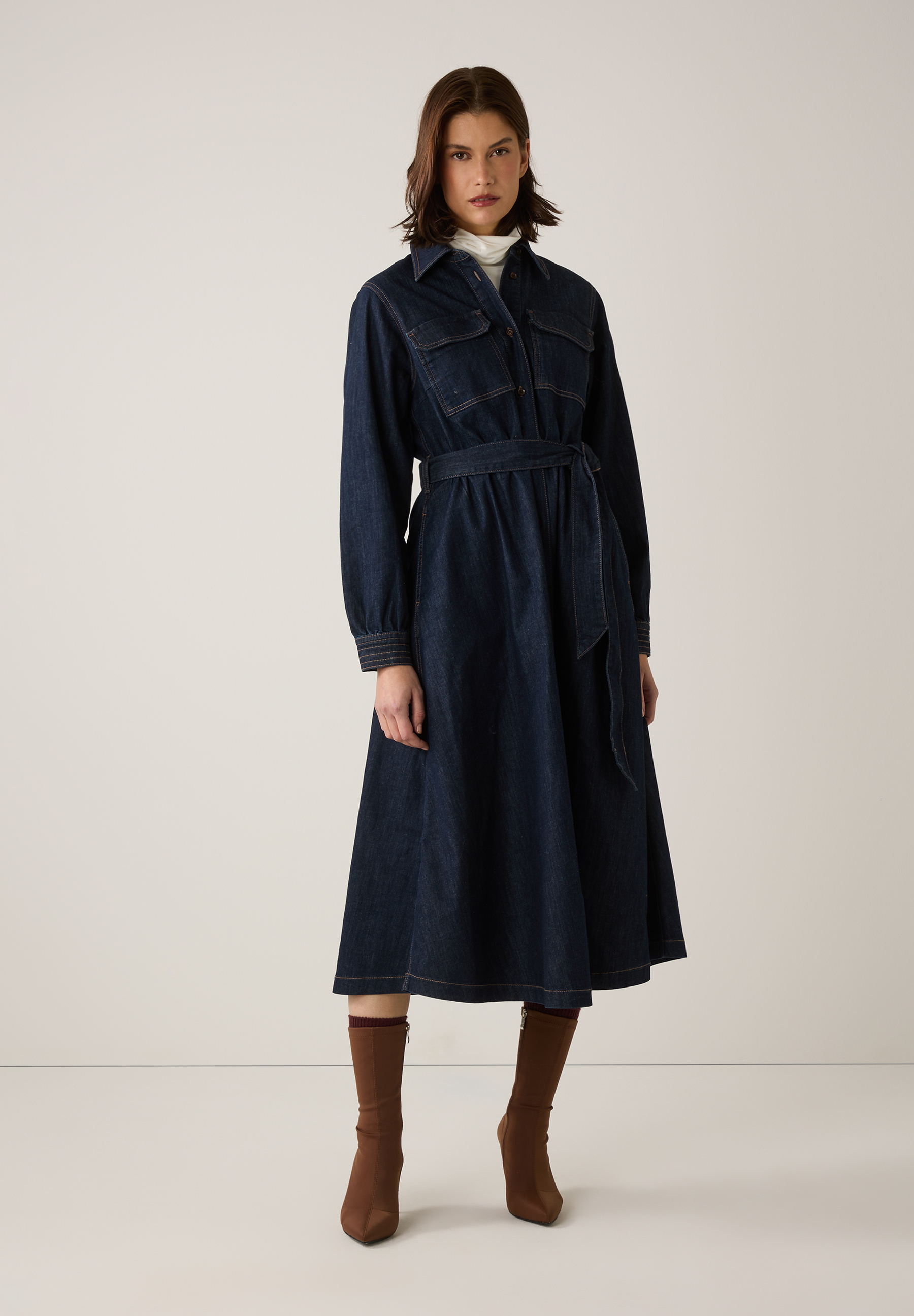 Eine Frau mit einem MORE & MORE-Jeanskleid mit langen Ärmeln und Gürtel, dazu braune Stiefel.