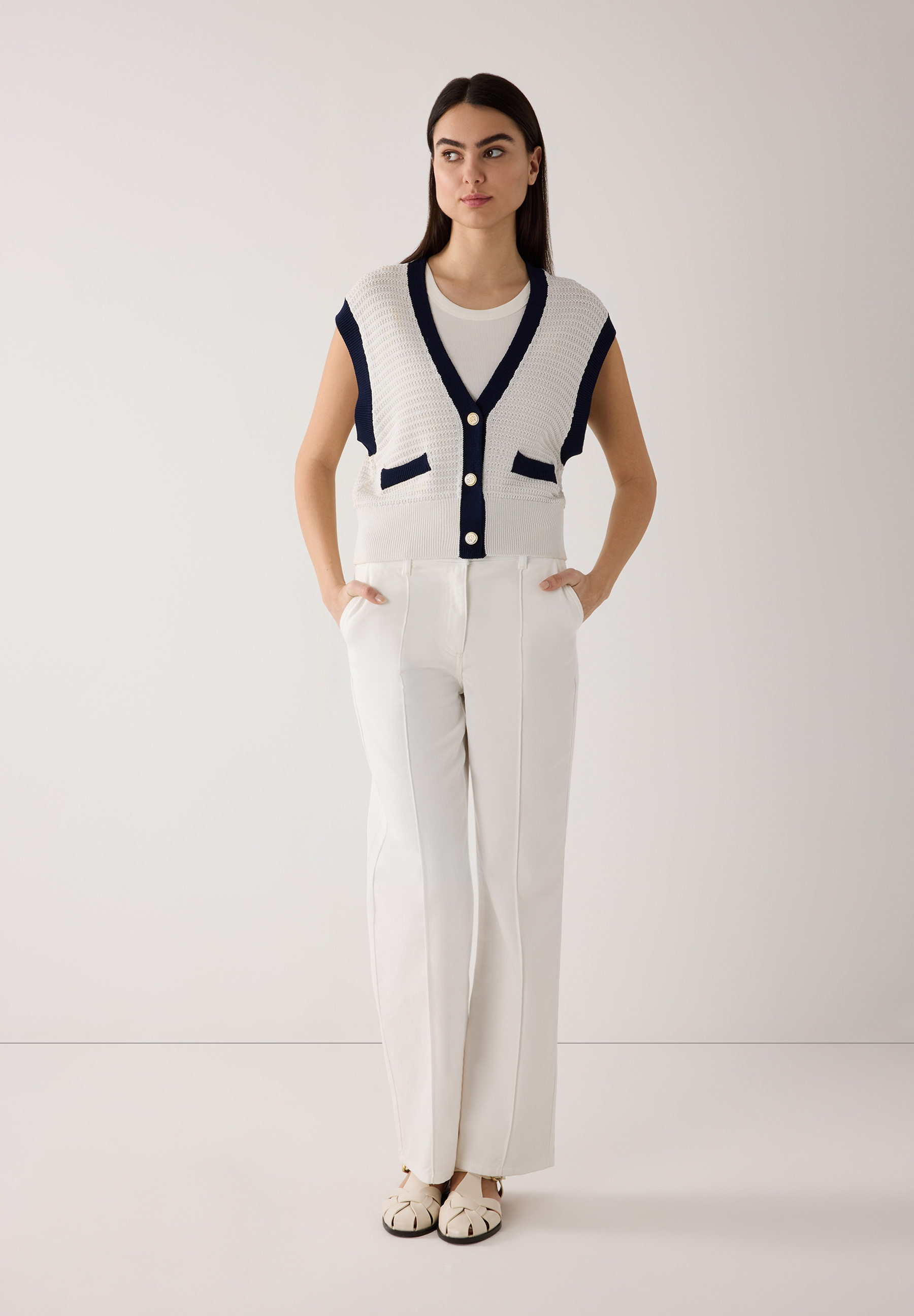 Eine Frau in MORE & MORE Strickware: Strukturstrick-Weste mit V-Ausschnitt, weißen Hosen und beigen Schuhen vor weißem Hintergrund. Eine Frau in MORE & MORE Strickware: Strukturstrick-Weste mit V-Ausschnitt, weißen Hosen und beigen Schuhen vor weißem Hintergrund.