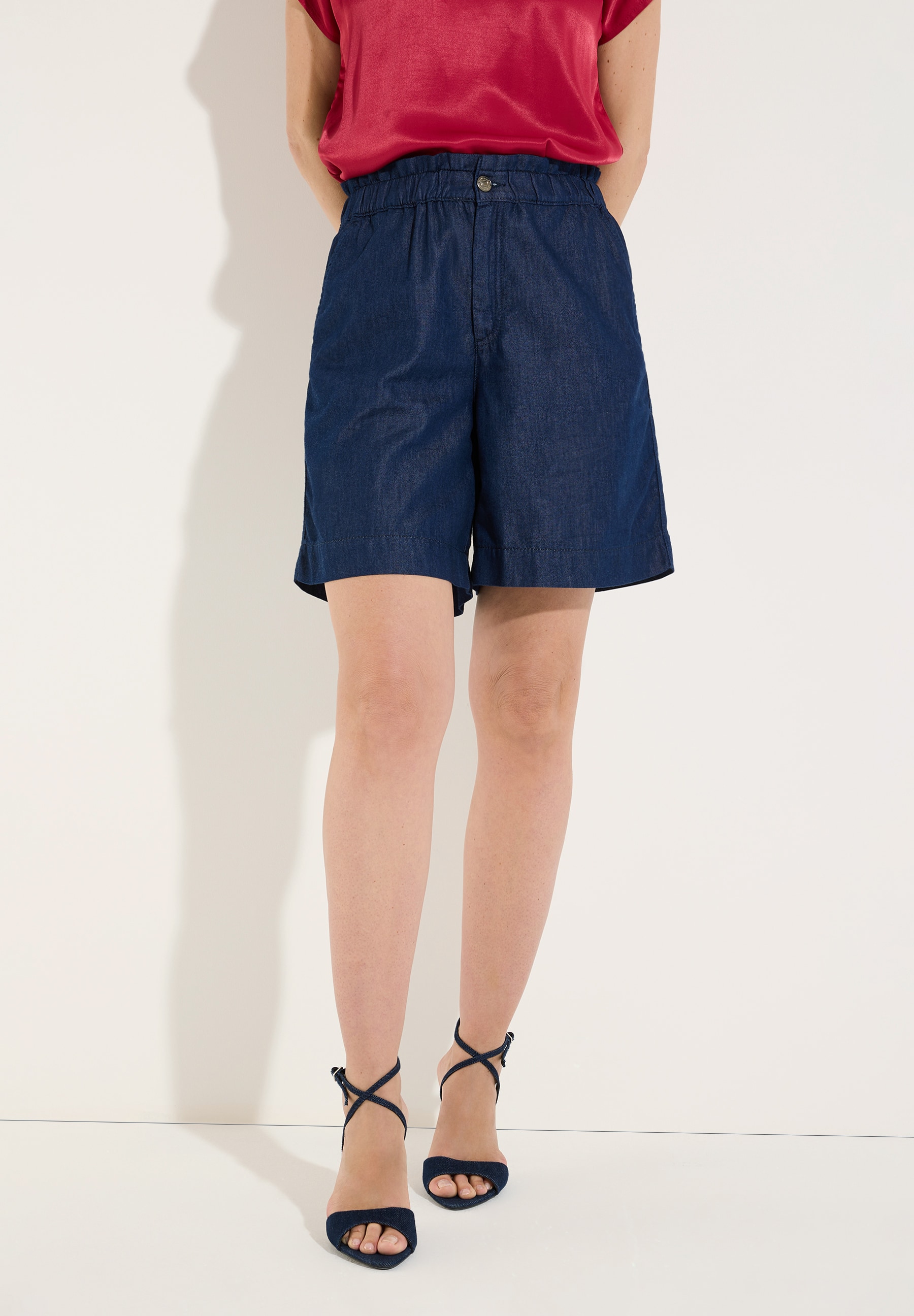 MORE & MORE Jeans-Shorts aus angenehmem Denim in Dunkelblau, kombiniert mit einem roten Oberteil und hohen Sandalen.