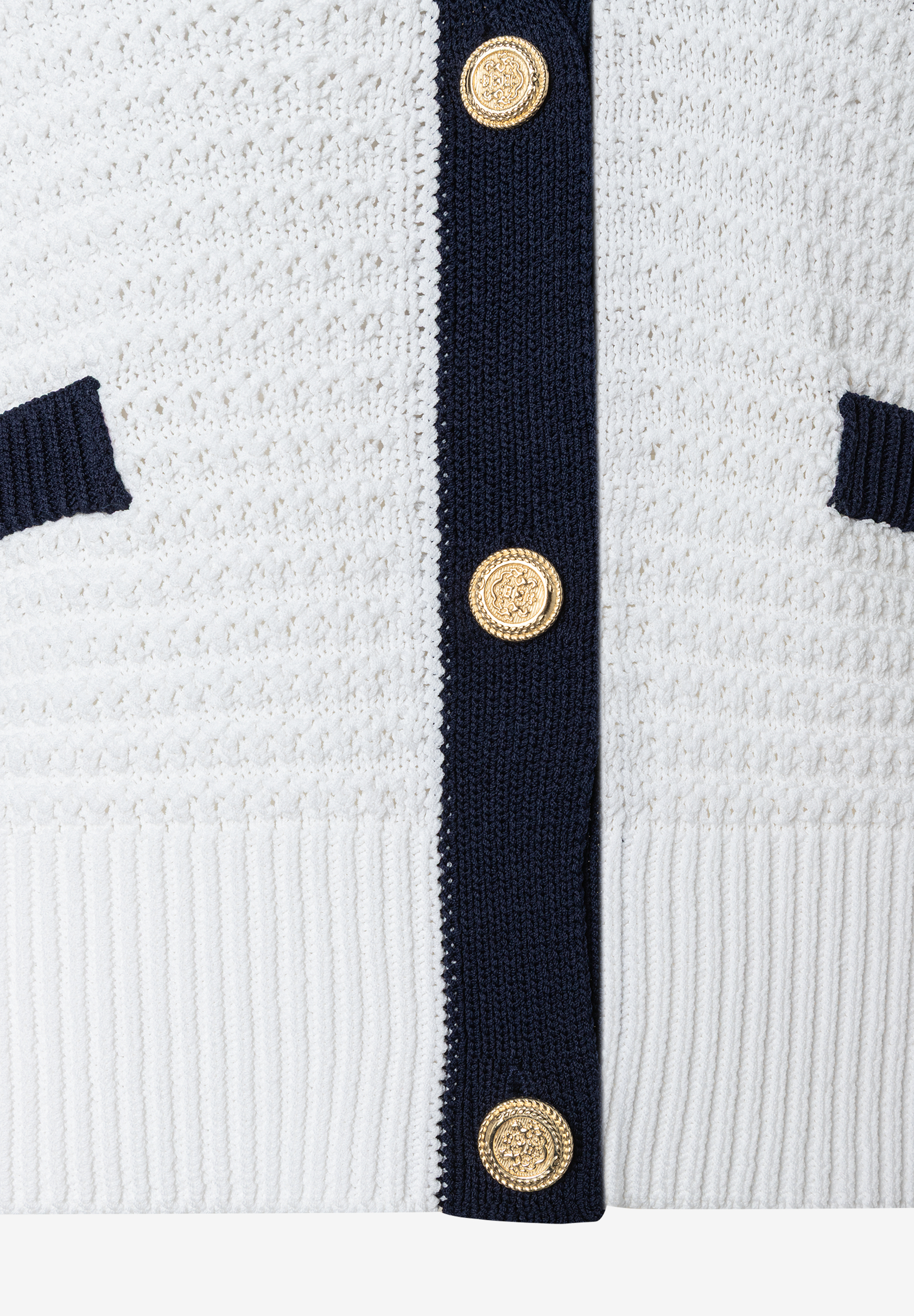 Nahaufnahme eines weißen MORE & MORE Strickoberteils mit blauen Akzenten und goldenen Knöpfen. Nahaufnahme eines weißen MORE & MORE Strickoberteils mit blauen Akzenten und goldenen Knöpfen.