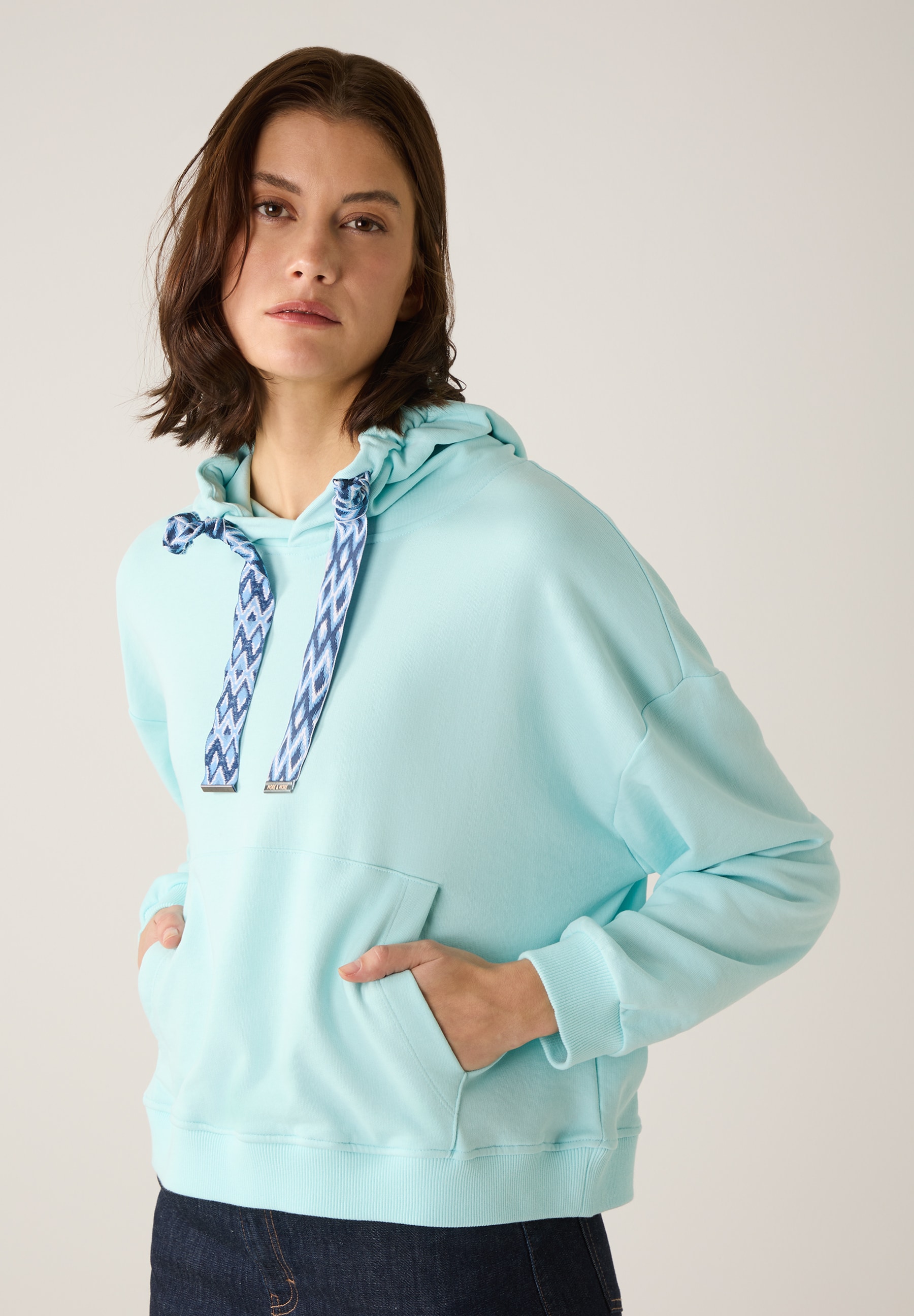 Eine Frau mit einem MORE & MORE Sweatshirt mit Kapuze in einem hellen Türkiston, die Hand in der Tasche.