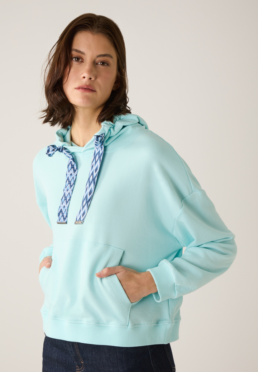 Eine Frau mit einem MORE & MORE Sweatshirt mit Kapuze in einem hellen Türkiston, die Hand in der Tasche. Eine Frau mit einem MORE & MORE Sweatshirt mit Kapuze in einem hellen Türkiston, die Hand in der Tasche.