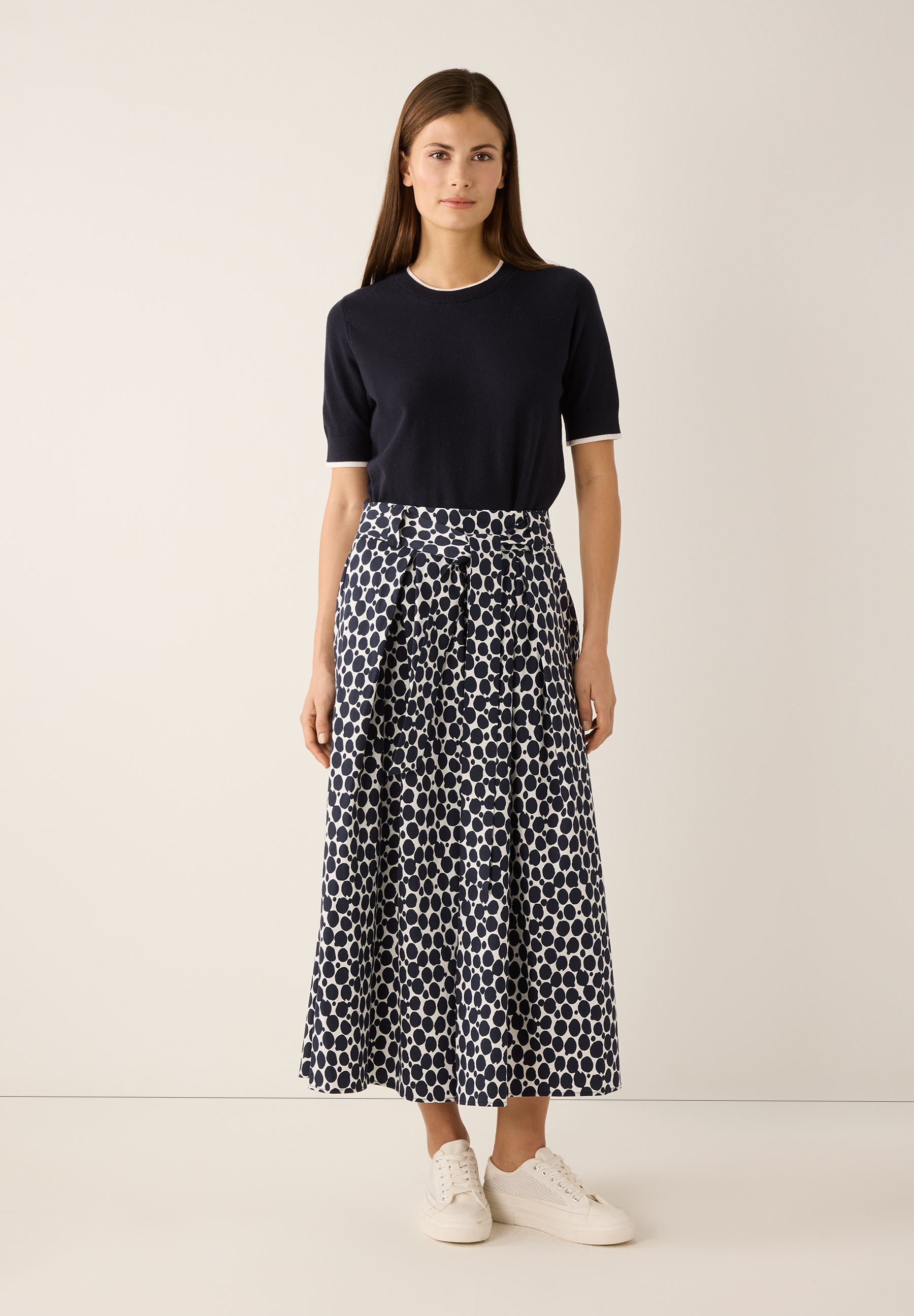 Eine Frau in MORE & MORE Feinstrick-Oberteil und einem Rock mit Punktmuster, dazu weiße Sneaker. Eine Frau in MORE & MORE Feinstrick-Oberteil und einem Rock mit Punktmuster, dazu weiße Sneaker.
