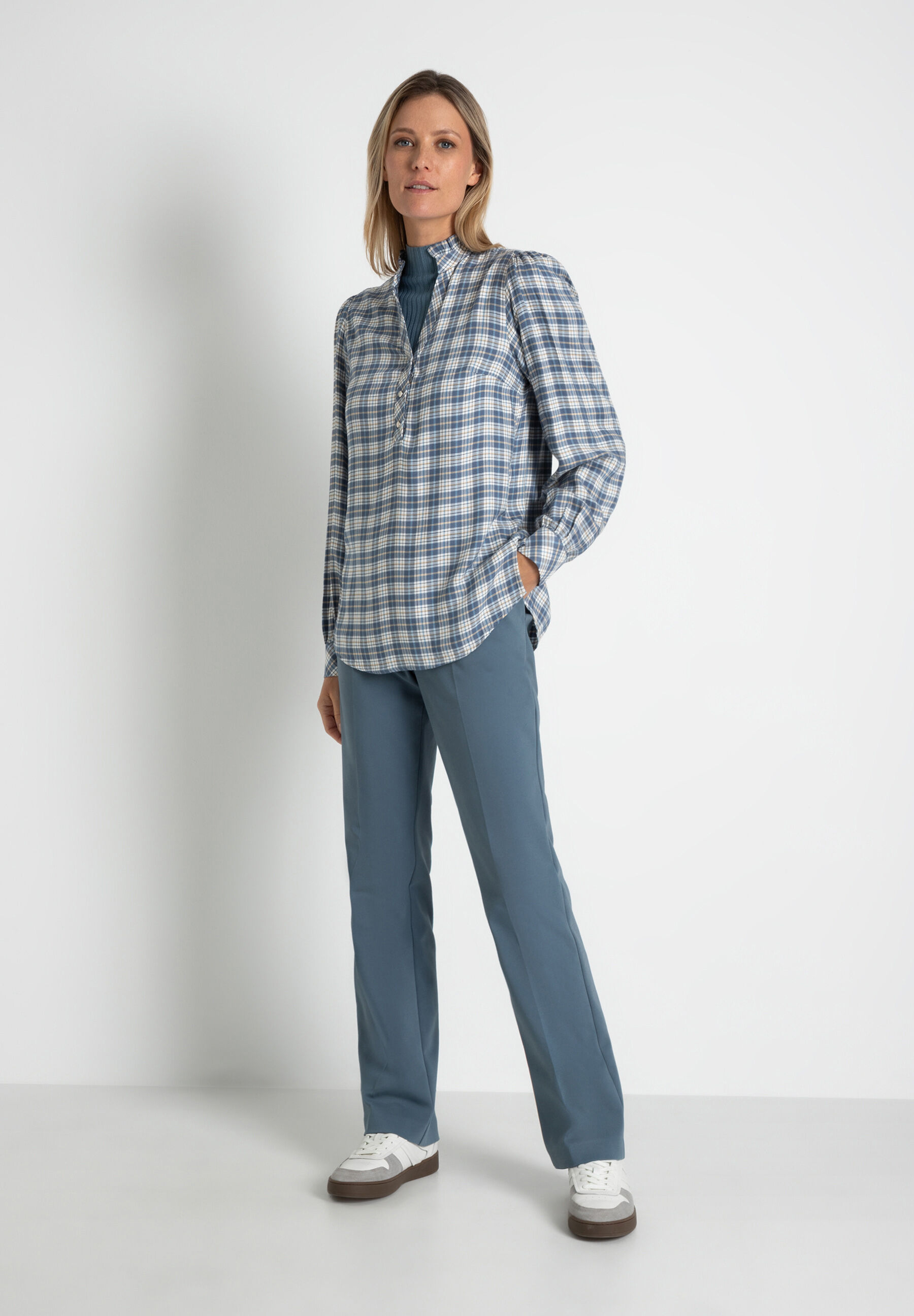 Eine Frau in MORE & MORE Oberteilen, karierter Bluse und passender Hose vor weißem Hintergrund. Eine Frau in MORE & MORE Oberteilen, karierter Bluse und passender Hose vor weißem Hintergrund.