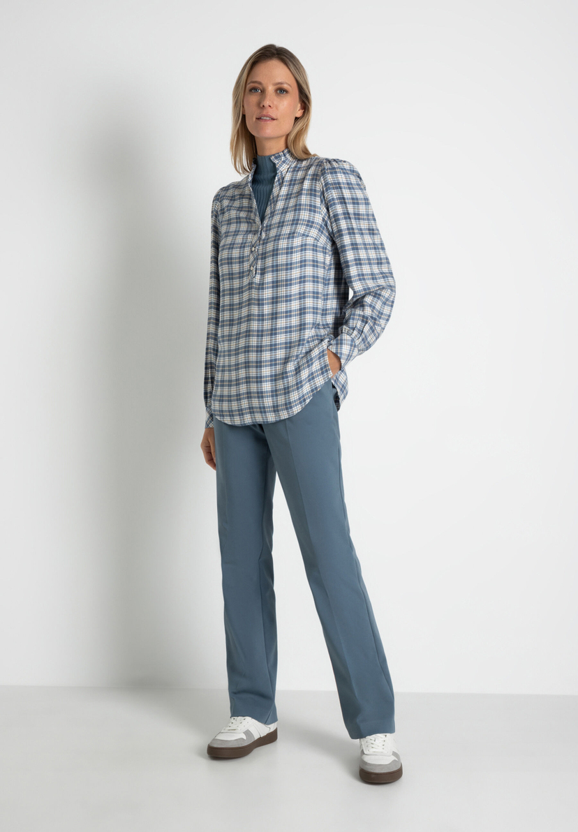 Eine Frau in MORE & MORE Oberteilen, karierter Bluse und passender Hose vor weißem Hintergrund. Eine Frau in MORE & MORE Oberteilen, karierter Bluse und passender Hose vor weißem Hintergrund.