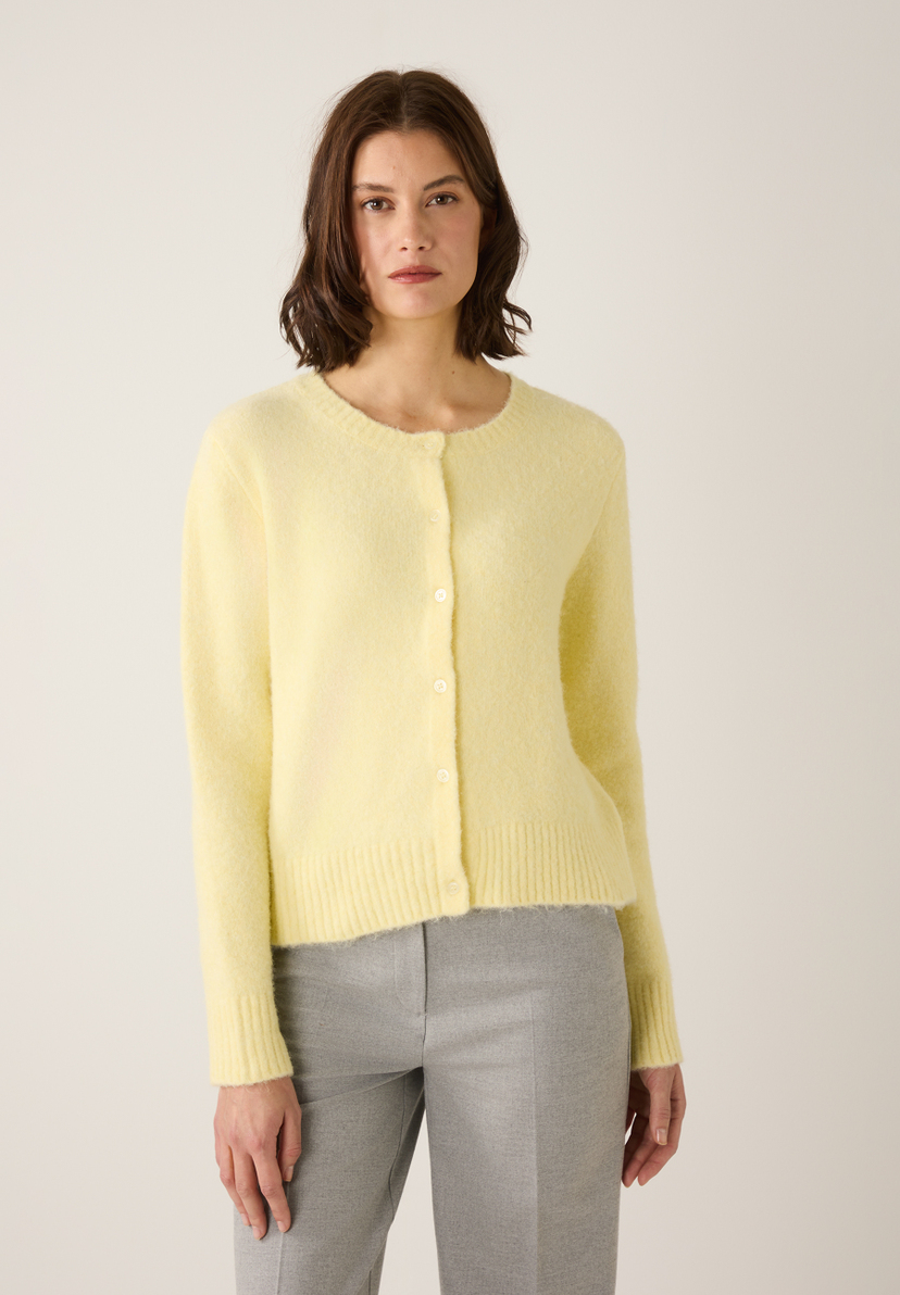Eine Frau in einem gelben MORE & MORE Strickoberteil mit Knöpfen und grauer Hose vor hellem Hintergrund. Eine Frau in einem gelben MORE & MORE Strickoberteil mit Knöpfen und grauer Hose vor hellem Hintergrund.