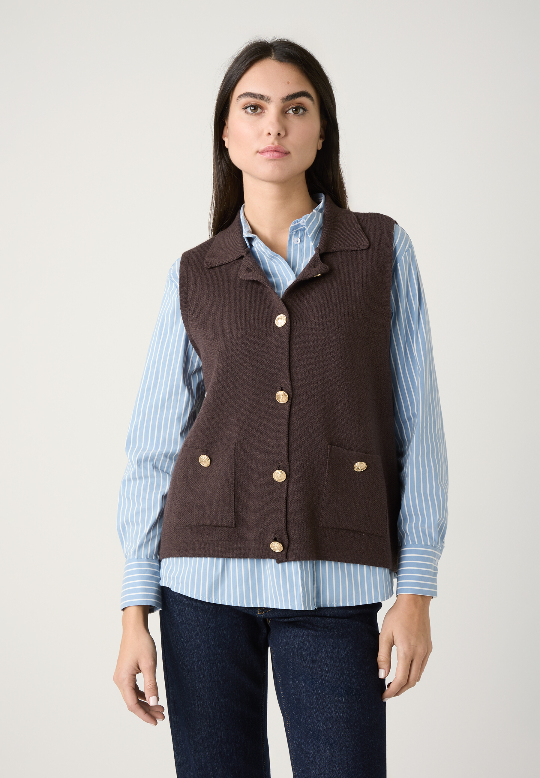 Eine Frau in einem MORE & MORE Strickoberteil mit goldenen Knöpfen, darunter eine blau-weiß gestreifte Bluse und Jeans.