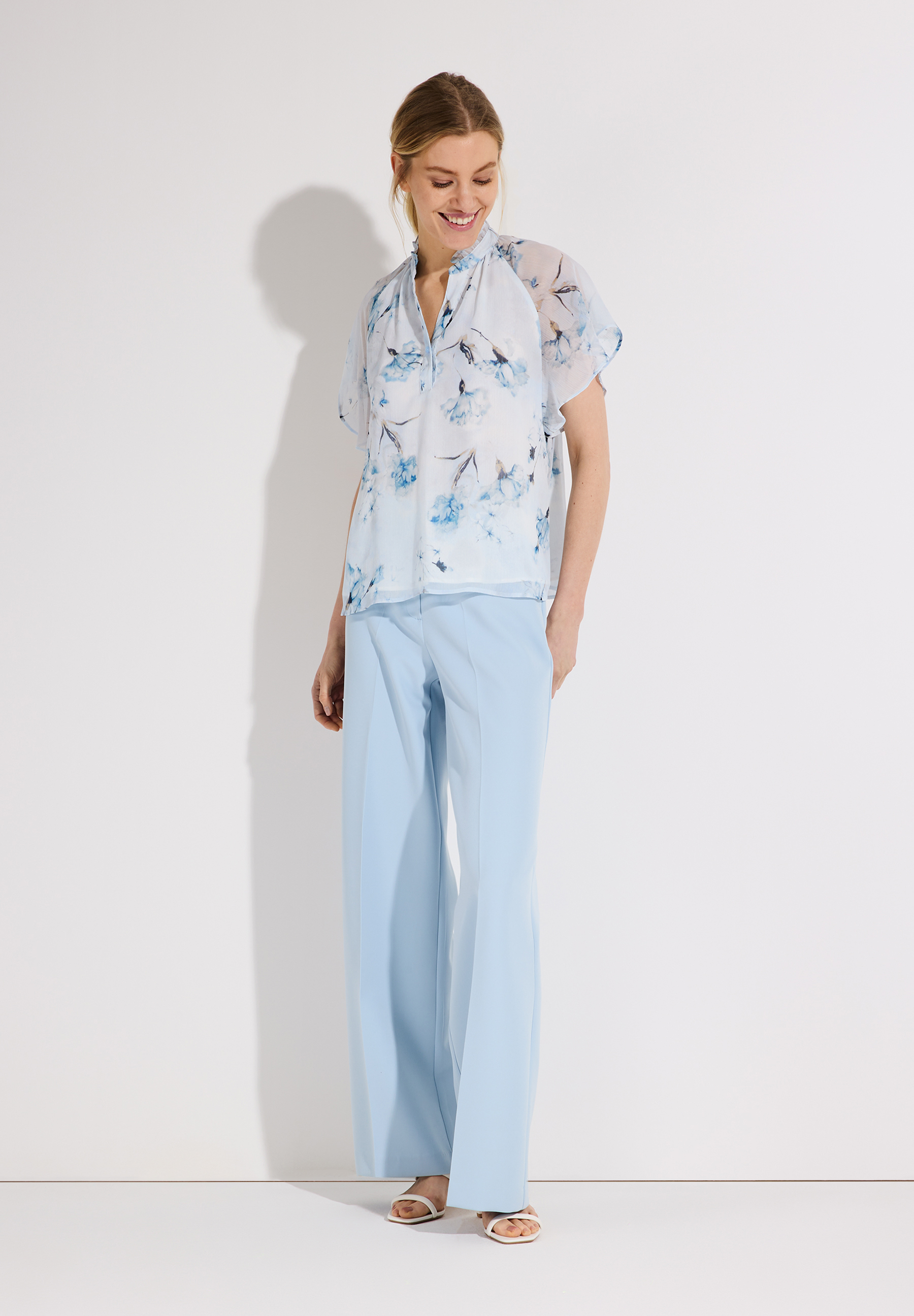 Eine Frau in MORE & MORE Chiffon-Bluse mit Blumenmuster und hellblauer Hose.