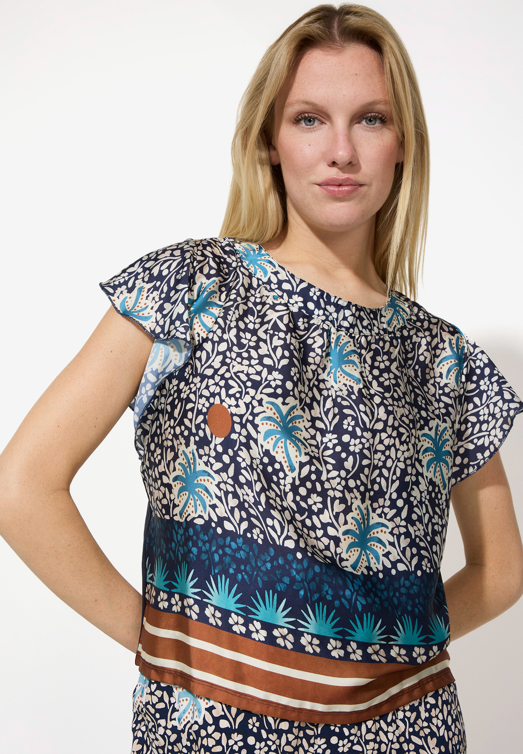 Eine Frau mit einem MORE & MORE Top mit blau-weißem Blumenmuster posiert vor einem weißen Hintergrund. Eine Frau mit einem MORE & MORE Top mit blau-weißem Blumenmuster posiert vor einem weißen Hintergrund.