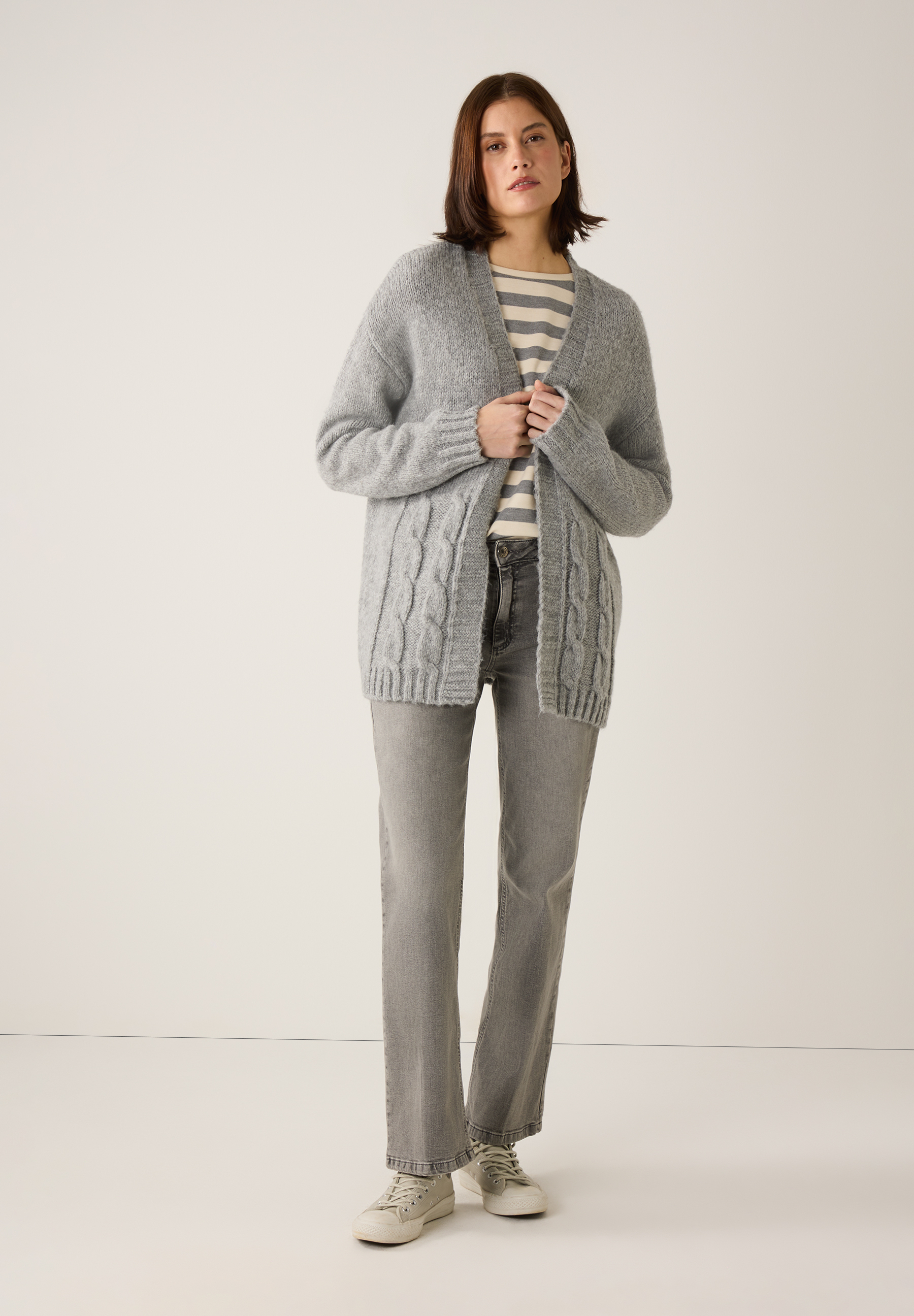 Eine Frau in MORE & MORE Oberteilen: graue Strickjacke, Jersey-Top, Jeans und Turnschuhe vor hellem Hintergrund. Eine Frau in MORE & MORE Oberteilen: graue Strickjacke, Jersey-Top, Jeans und Turnschuhe vor hellem Hintergrund.