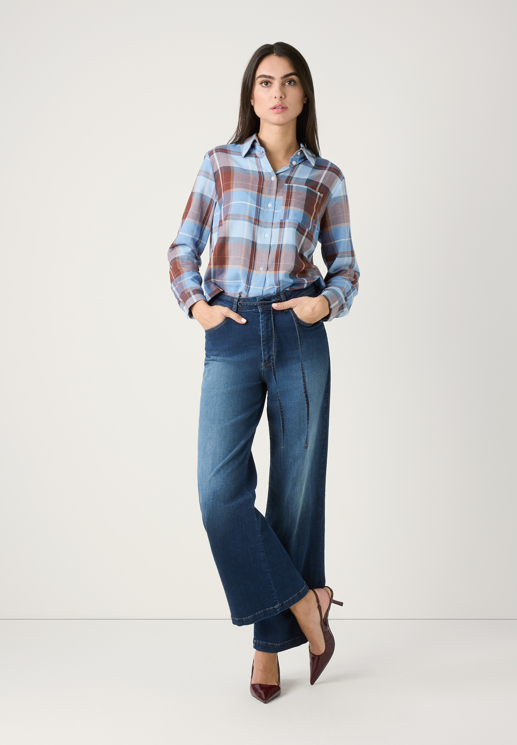 Eine Frau, die MORE & MORE Jeans aus Denim und ein kariertes Hemd trägt, steht vor einem weißen Hintergrund. Eine Frau, die MORE & MORE Jeans aus Denim und ein kariertes Hemd trägt, steht vor einem weißen Hintergrund.