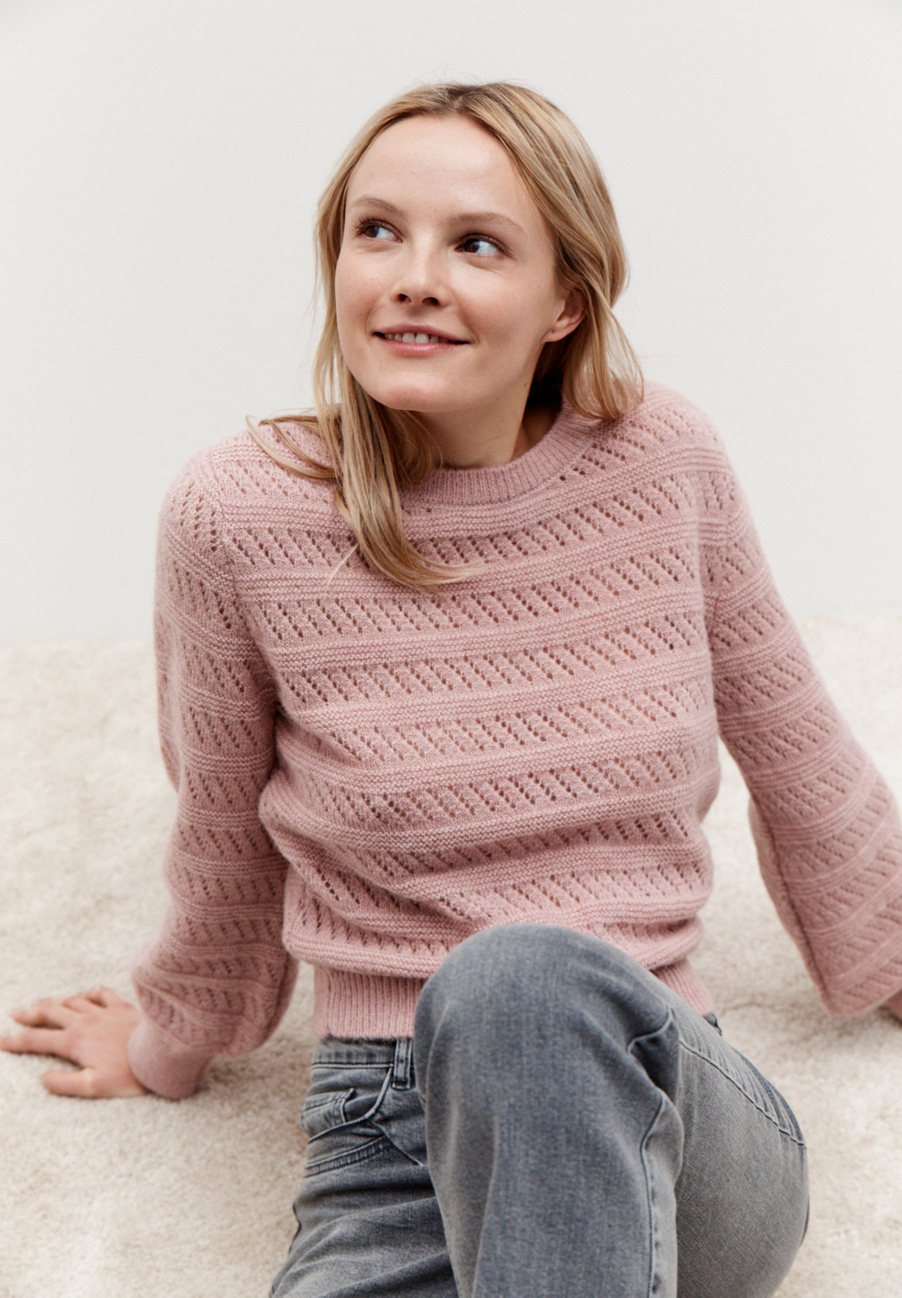 Eine Frau mit einem MORE & MORE Strukturstrick-Pullover sitzt auf einem Teppich und schaut lächelnd zur Seite.